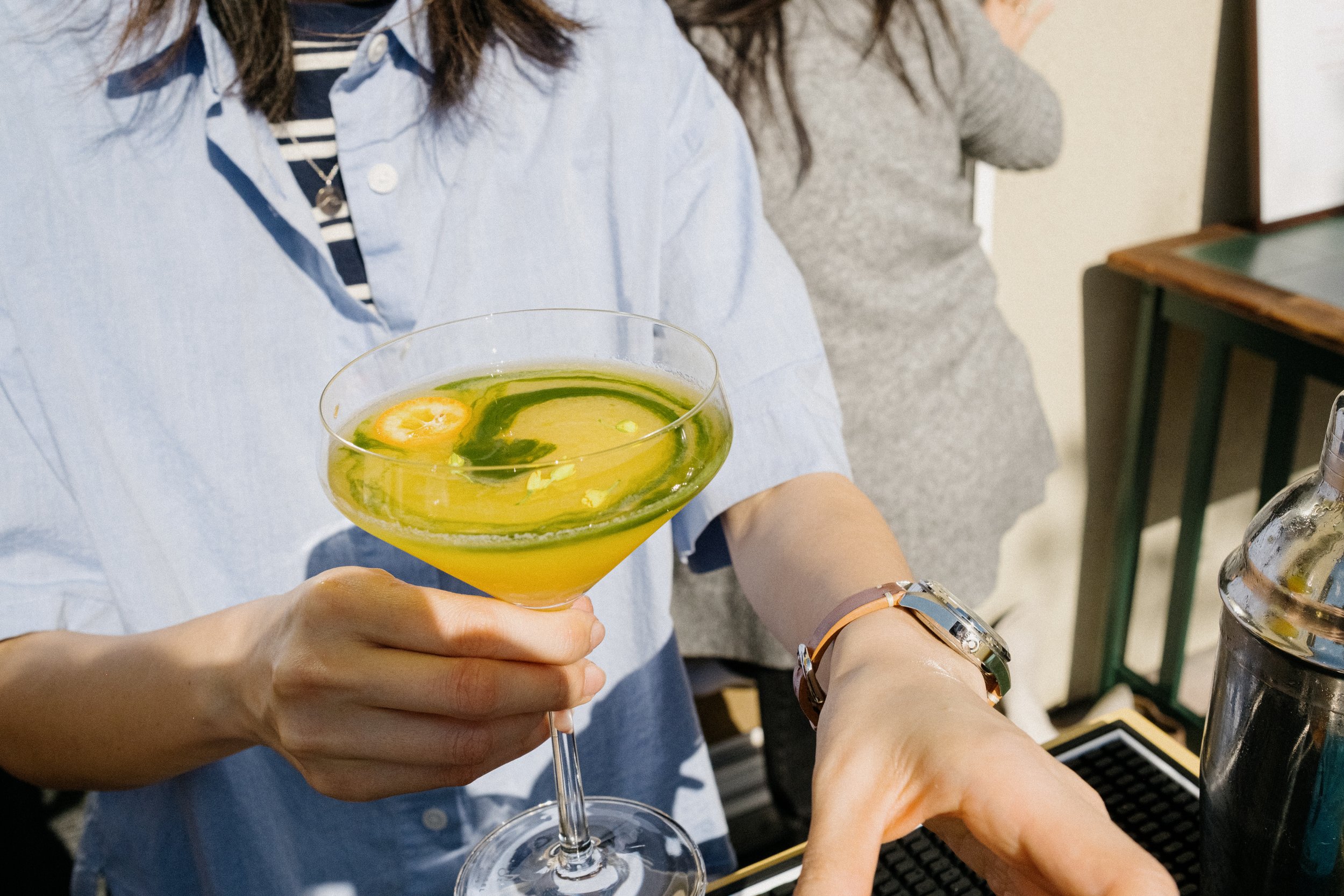 Person holding a kumquat honey mocktail with green matcha swirl garnish in a martini glass, wearing a denim shirt and watch, at a local pop up.