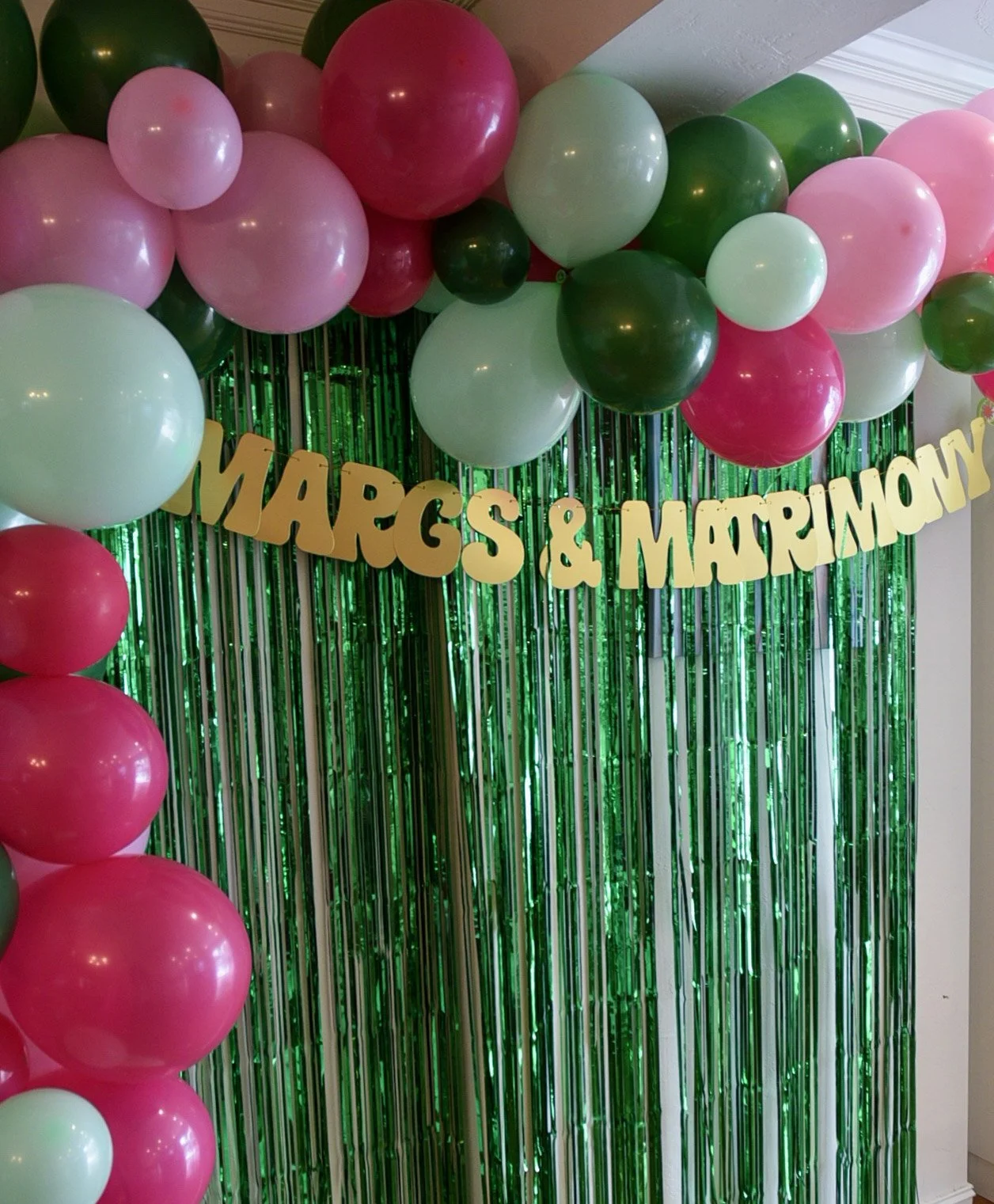 Decorative backdrop with pink, green, and white balloons, gold letter banner saying 'Margs & Matrimony,' and green metallic fringe curtain.