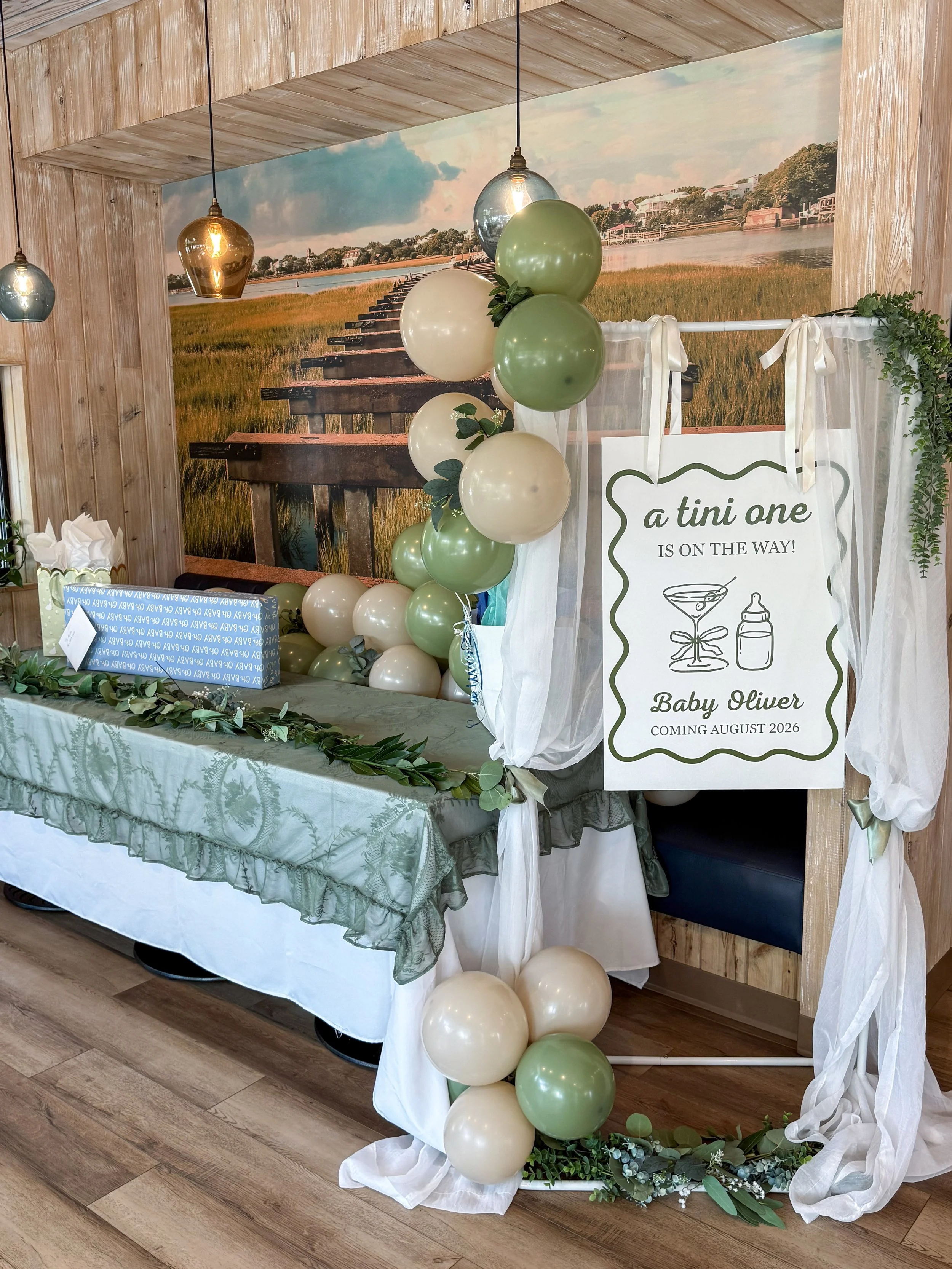 Baby shower decoration with a backdrop of a wooden pier over water, green and cream balloons, a sign that says 'a tiny one is on the way! Baby Oliver coming August 2026', greenery, and white sheer curtains.