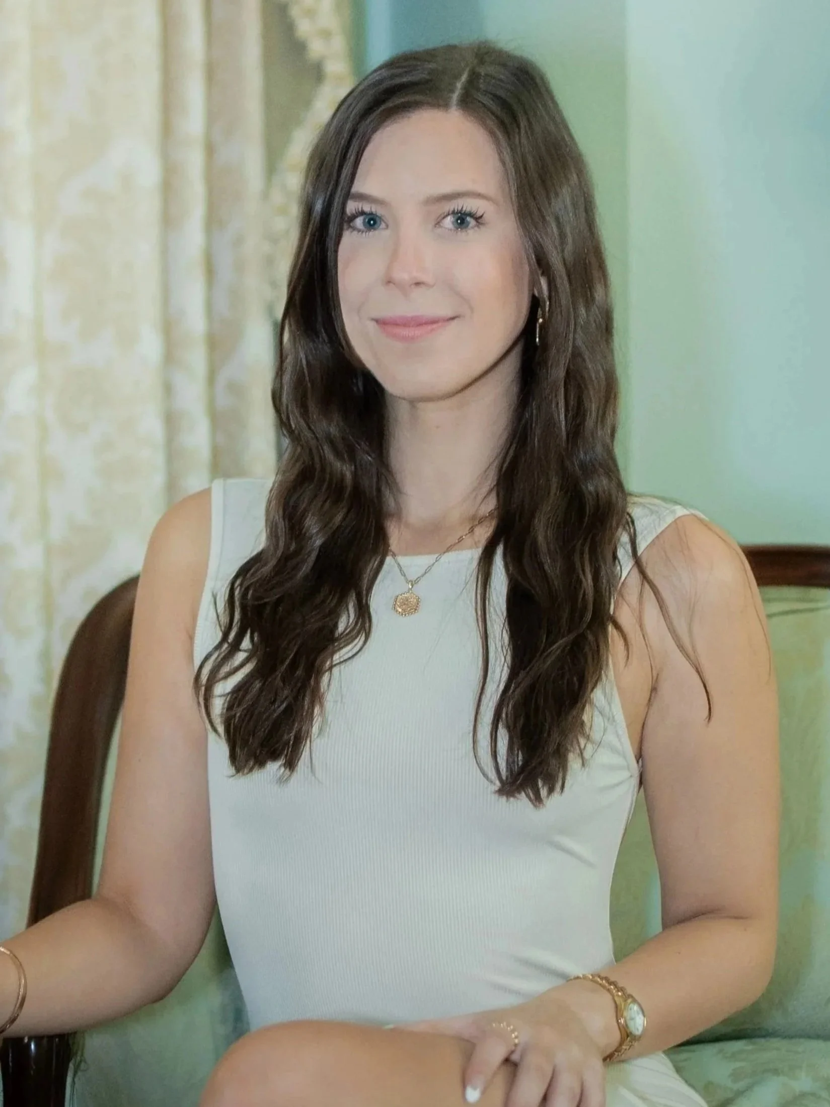 A young woman with long, wavy brown hair and bright blue eyes, smiling softly, sitting at a table. She is wearing a white sleeveless top, gold jewelry including a watch, bracelet, and earrings, and is seated indoors with a green wall and patterned curtains in the background.