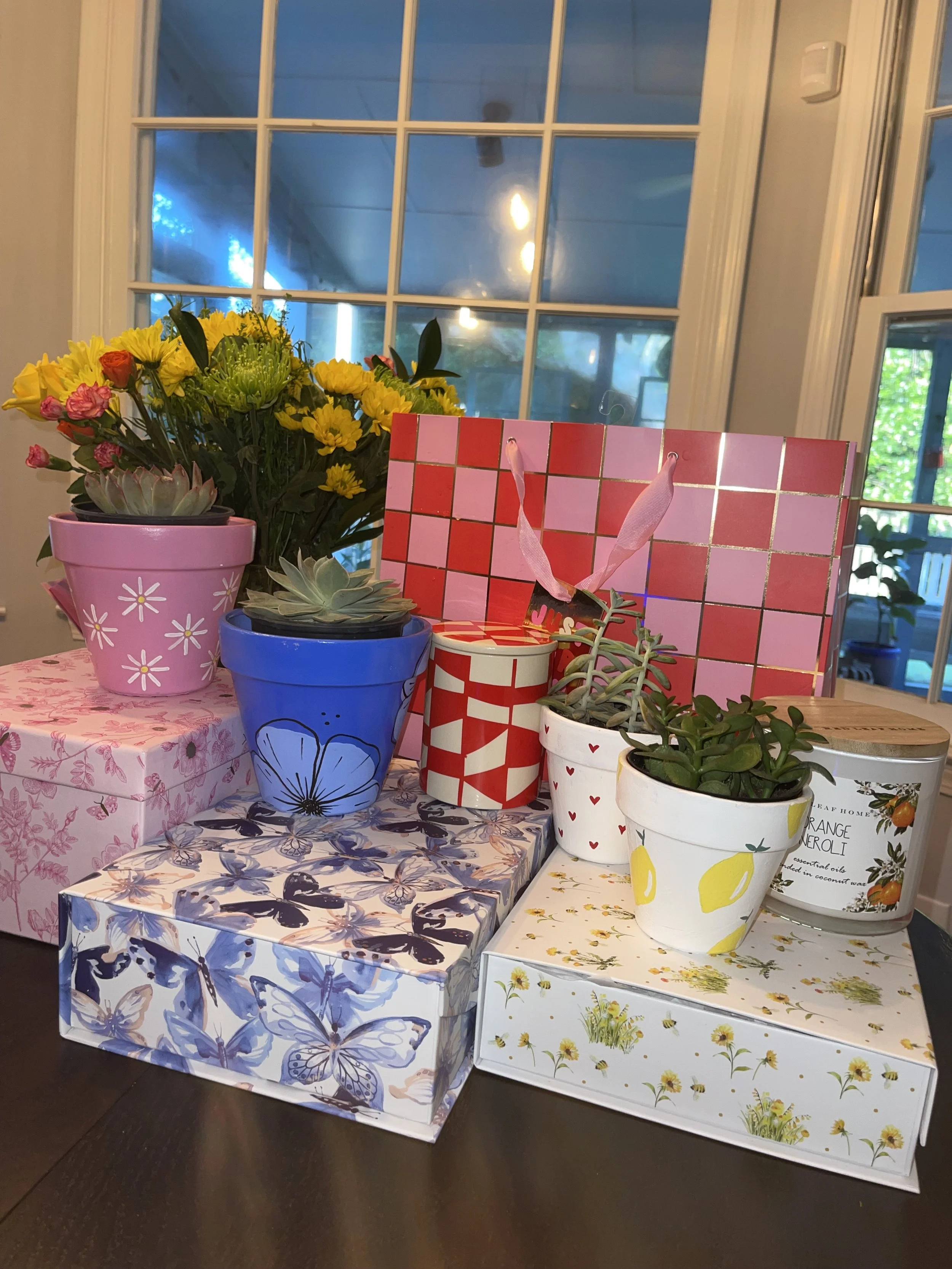 A collection of colorful flower pots and boxes on a table near a window. The pots include a pink pot with yellow flowers, a blue pot with succulents, and a white pot with green plants. There are decorative boxes with floral and butterfly patterns. 