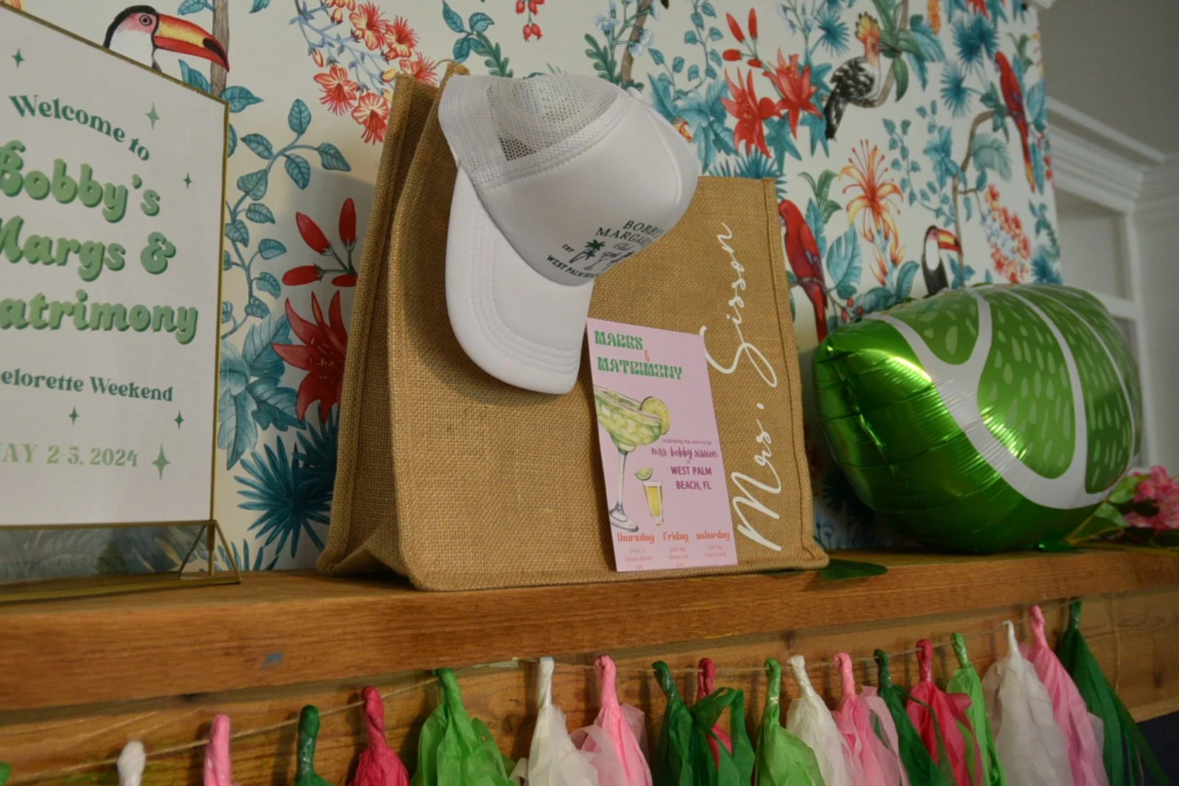 A colorful tropical-themed background on a wooden surface, a partially visible white cap, a tan bag, a pink drink menu, a metallic green balloon shaped like a lime, and a small, colorful paper garland hanging on a string.