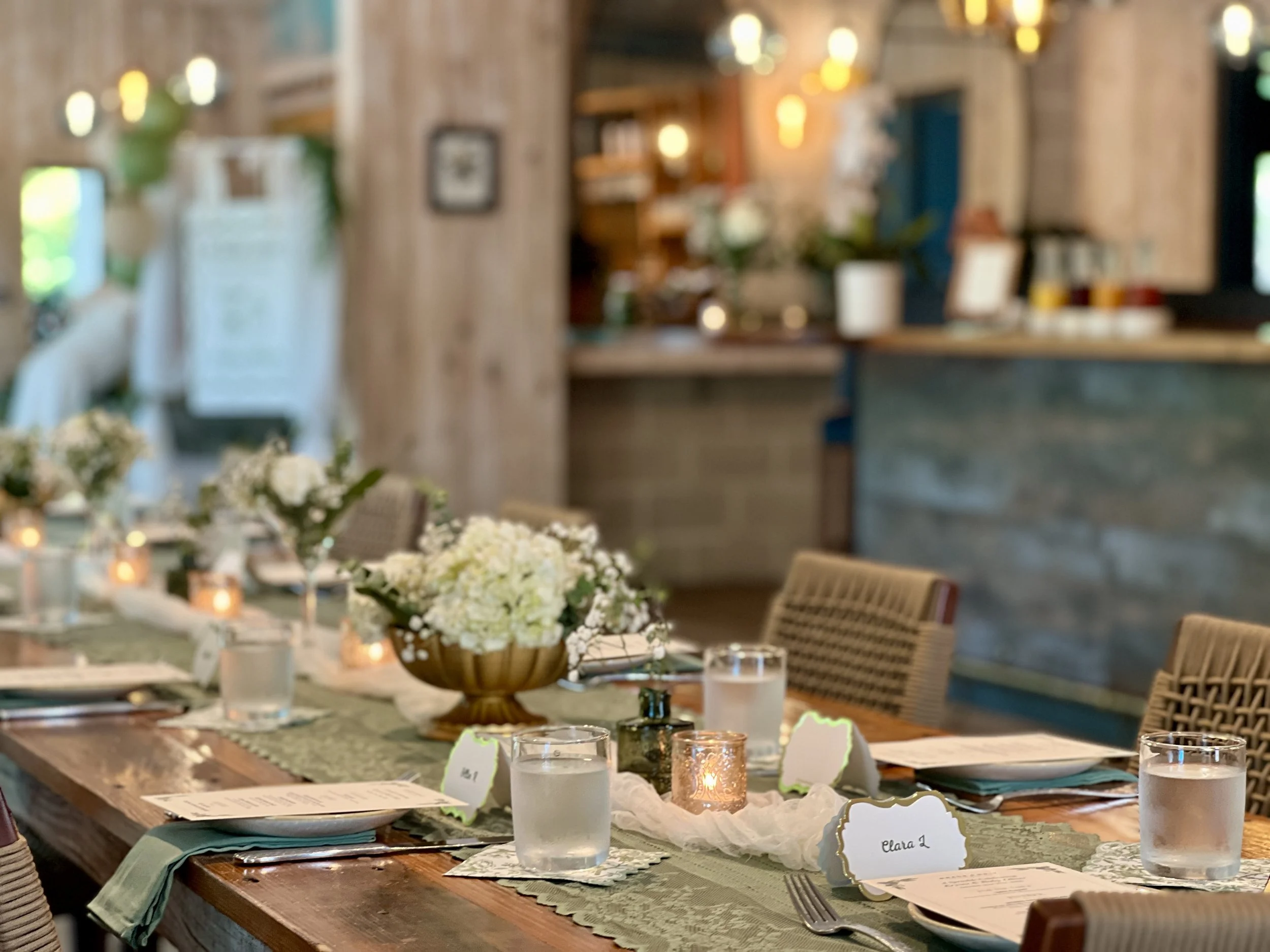 A close-up of a dining table set for a celebration with flower arrangements, candles, name cards, and glasses, in a rustic, warmly lit room.