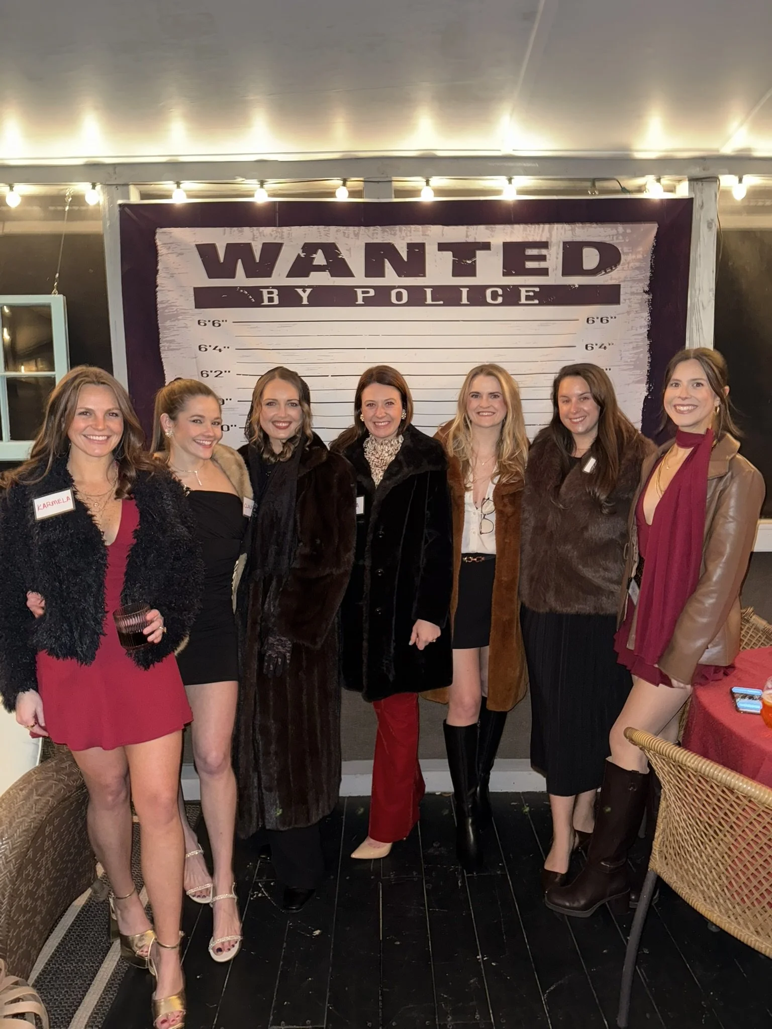 A group of six women standing together in front of a large wanted poster backdrop with height measurement markings, at a social event. They are smiling and dressed in stylish clothes.