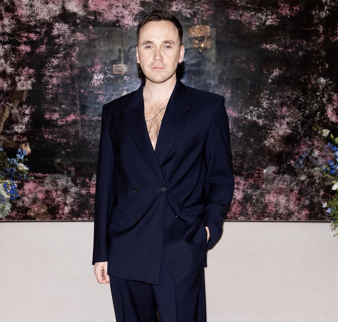 A man in a dark navy suit stands in front of a colorful abstract background with pink and purple hues, surrounded by floral decor.