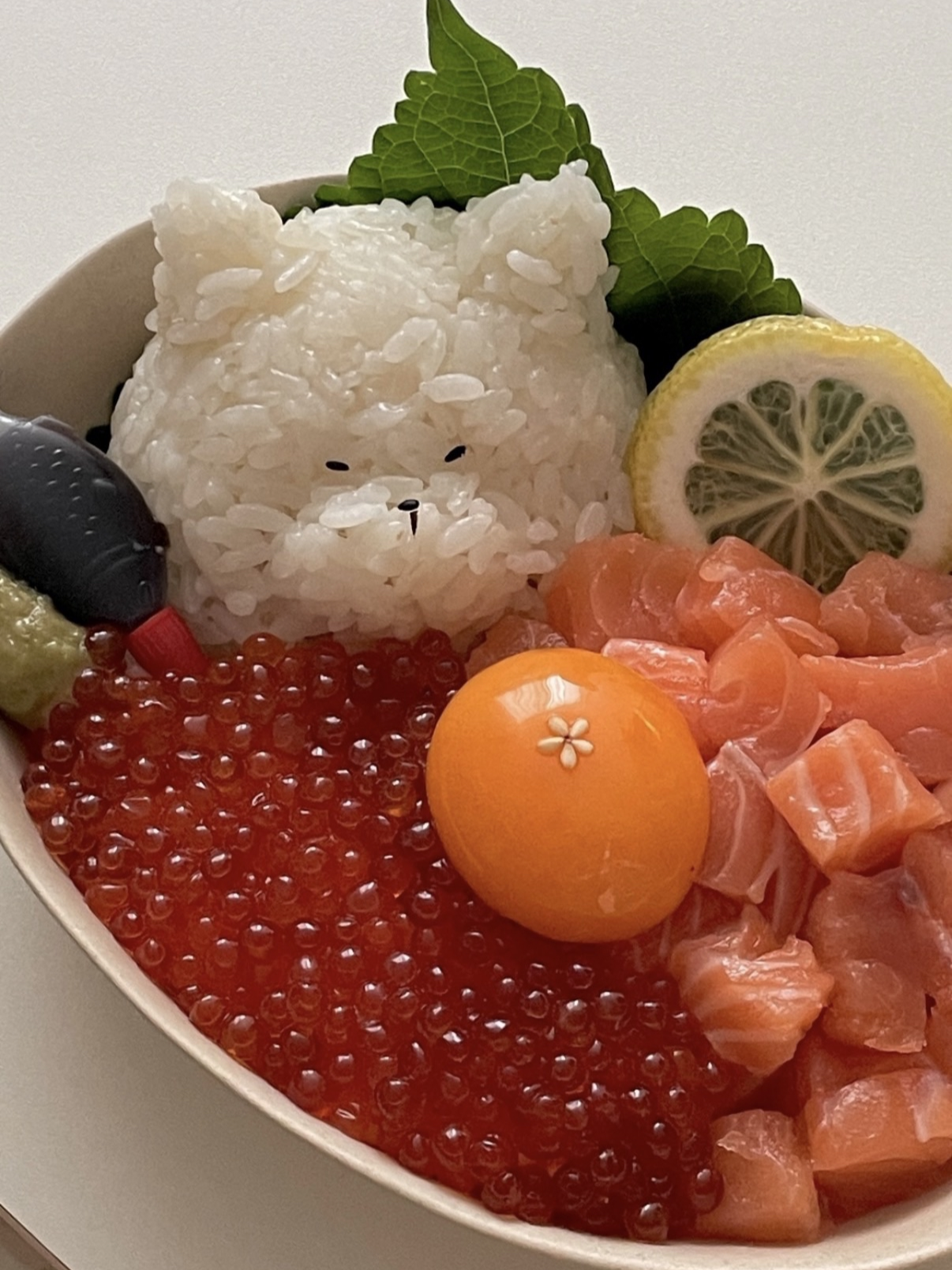 A bowl of sushi ingredients arranged to resemble a cute bear face, including rice for the face, salmon for the mouth, salmon roe for the cheeks, a lemon slice for the nose, and other garnishes like green leaves and decorative accents.