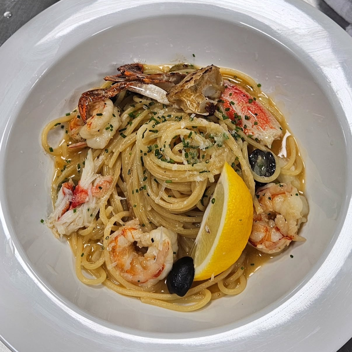 Seafood pasta with shrimp, crab claws, scallops, lemon wedge, black olives, and chopped herbs on a white plate.