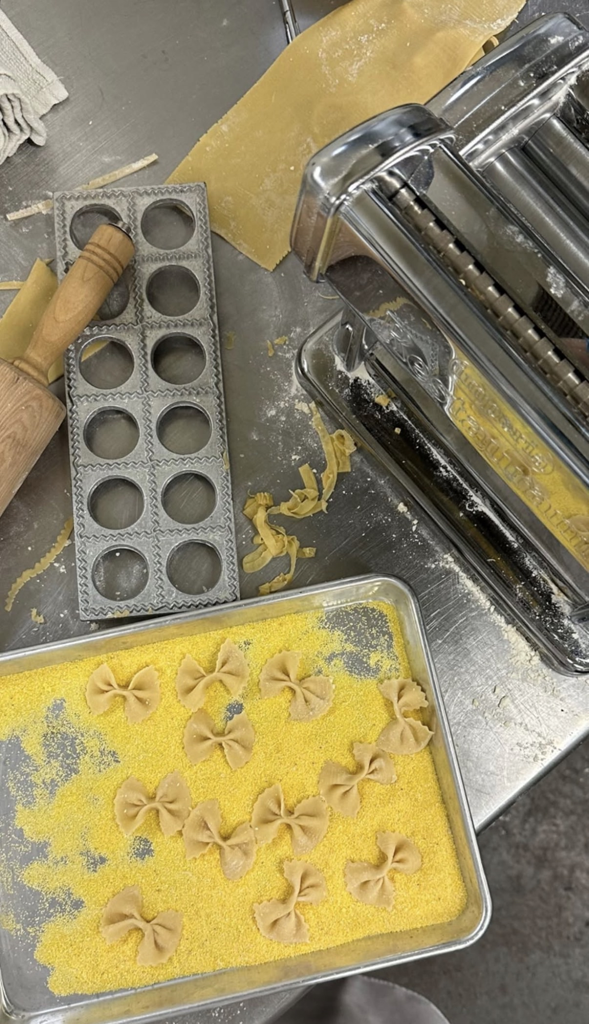 A tray of uncooked bowtie pasta on a yellow dish drying mat, with a pasta machine and dough rollers on a stainless steel countertop.