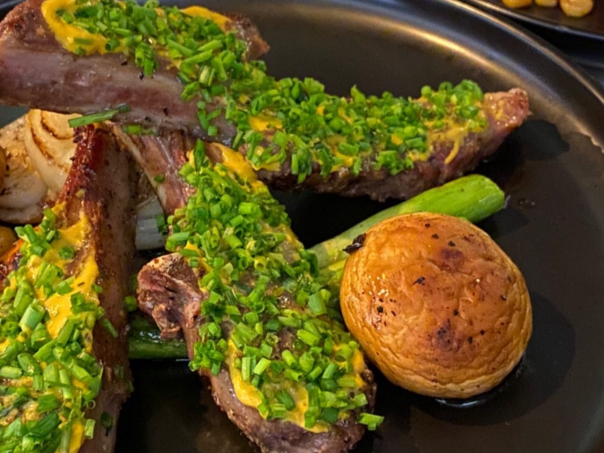 Cooked lamb chops topped with chopped green onions, accompanied by a roasted potato and a green onion stalk on a black plate.