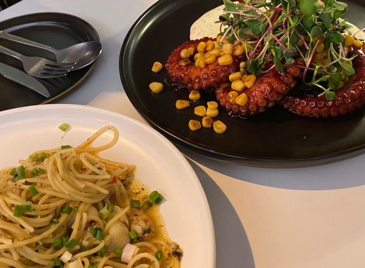A plate of spaghetti with chopped green onions and a side dish of cooked octopus tentacles topped with corn and microgreens.