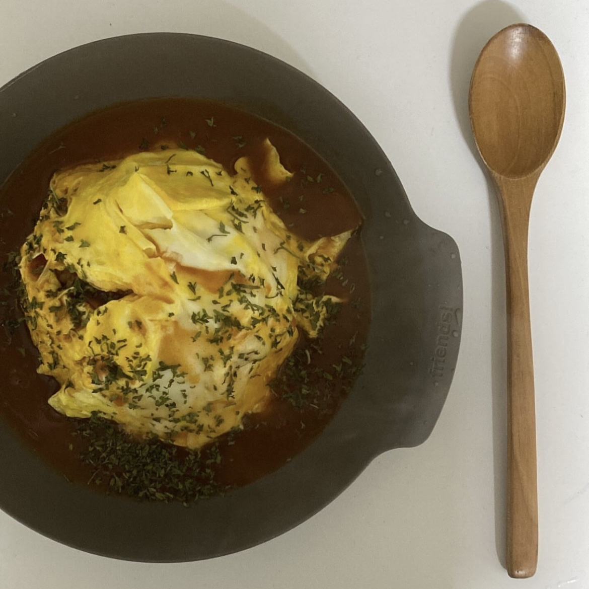 Omelette in a skillet with herbs, surrounded by sauce, with a wooden spoon on a white surface.