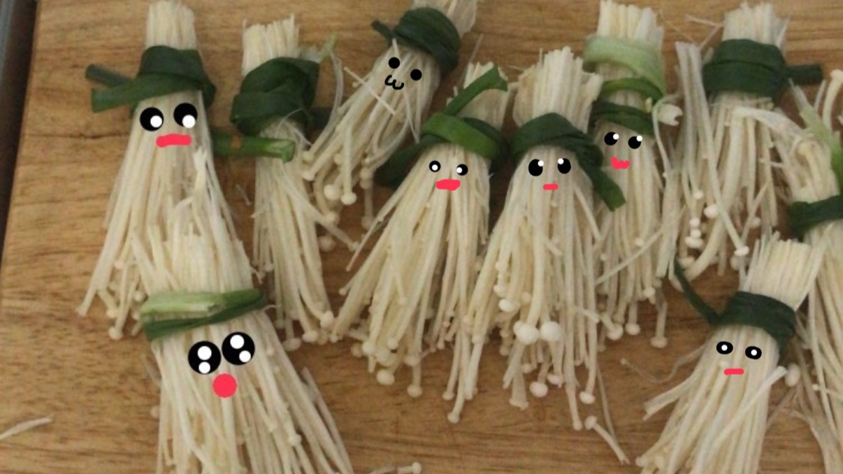 Cluster of enoki mushroom bundles decorated with green onion strips, each with cartoon faces drawn on, resembling tiny characters.