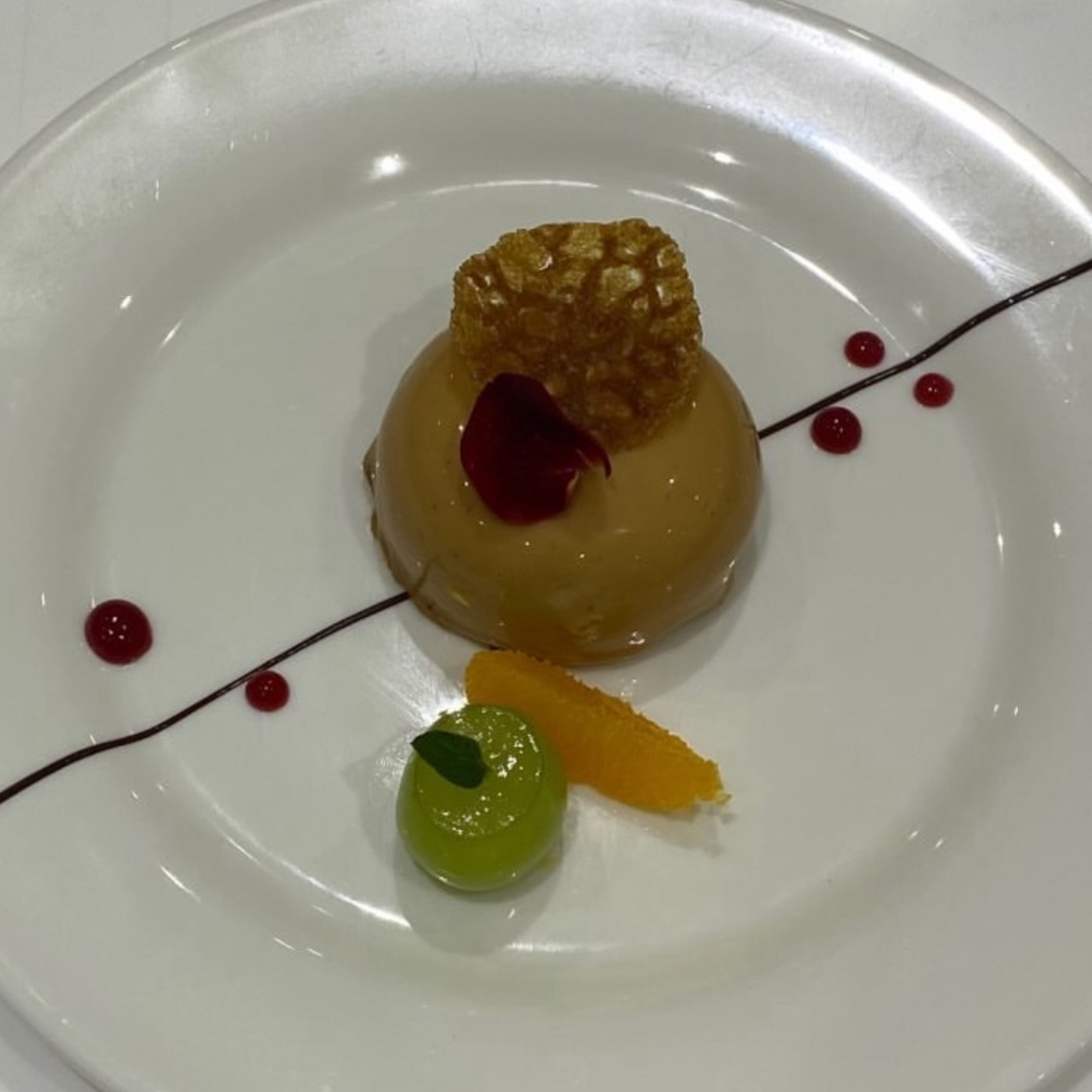A plated dessert featuring a round caramel or glazed pastry, topped with a small red petal and a textured, honey-colored piece of crisp. Garnished with a green grape, a slice of orange, and a small green leaf, with red sauce dots and a thin line of sauce on a white plate.