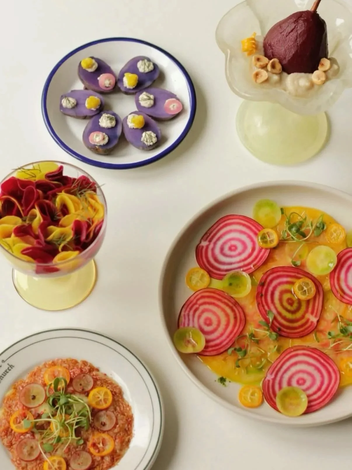 An assortment of colorful food dishes, including a purple floral-patterned plate of decorated cookies, a yellow and red pasta dish with vegetable toppings, a tart topped with small yellow and orange vegetables, and a fruit salad with sliced beets, grapes, and radishes.