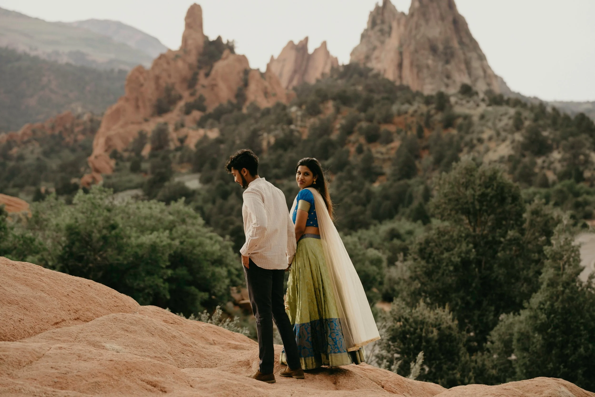 A man and woman standing on a rocky ledge outdoors, with a mountainous landscape and trees in the background. The woman is dressed in traditional Indian attire, a blue blouse with a beige and gold skirt, and a sheer dupatta. The man is wearing a white shirt and dark pants, looking down with his head slightly bowed. The woman is looking at the camera with a slight smile.