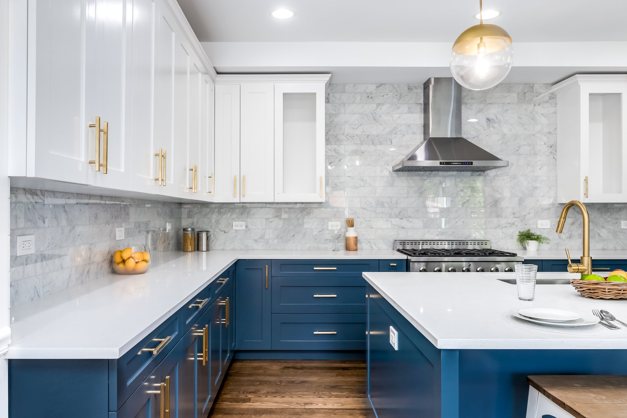 Modern kitchen with white marble backsplash, white upper cabinets, blue lower cabinets with gold handles, stainless steel range hood, and minimalist decor including a bowl of oranges, a glass of water, and a basket of green apples.