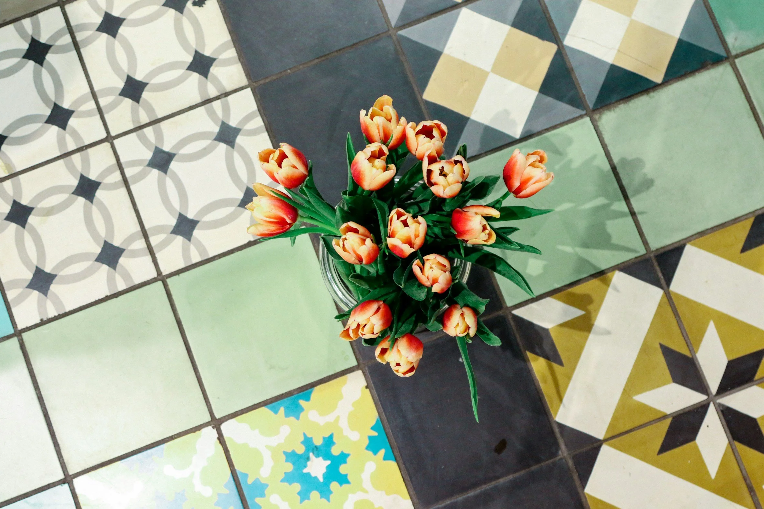 A bouquet of orange and yellow tulips in a glass vase on a colorful tiled floor with various geometric patterns.