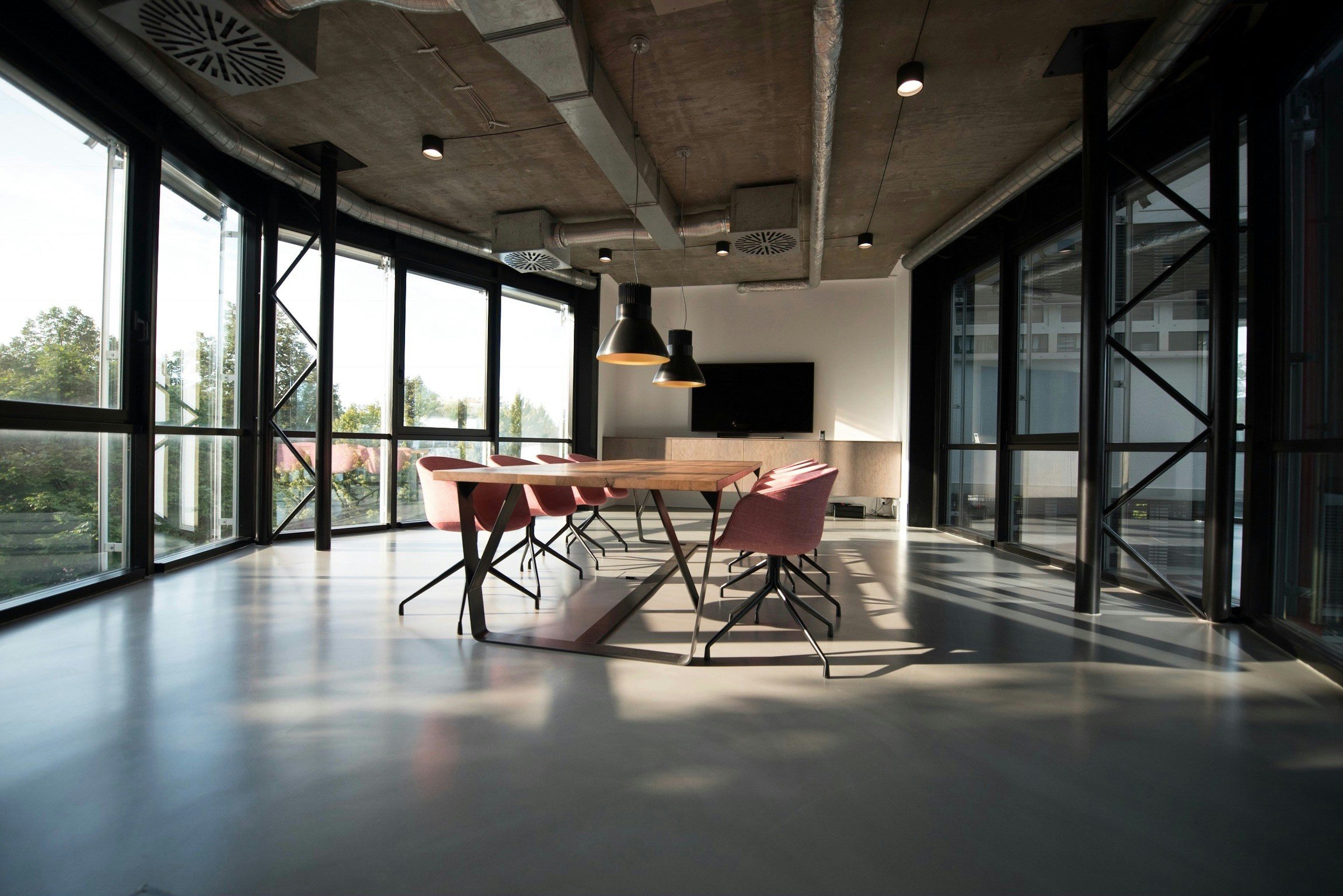 Modern office conference room with large floor-to-ceiling windows, a wooden table surrounded by pink chairs, ceiling-mounted pendant lights, a large flat-screen TV on a white wall, exposed ceiling ducts, and a view of trees outside.