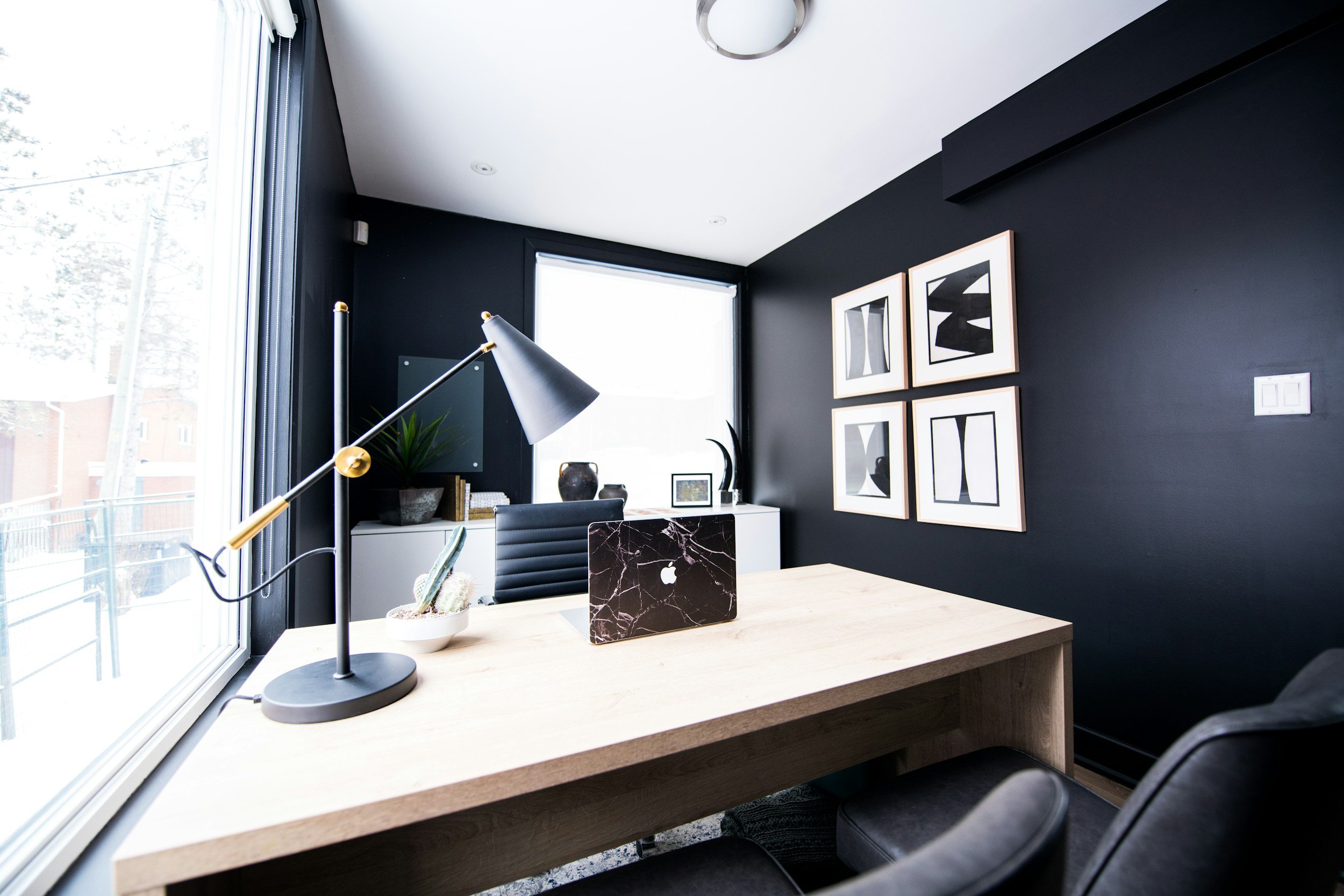 A modern home office with a light-colored wooden desk, a black office chair, and a marble laptop. The desk has a small cactus plant and a white pot. There is a large window on the left with a view of trees outside. The wall behind the desk is painted dark gray and features four framed abstract black-and-white art pieces. A black and white desk lamp is on the desk, and there are decorative items like vases and picture frames on a shelf near the window. The ceiling has recessed lighting and a round light fixture.