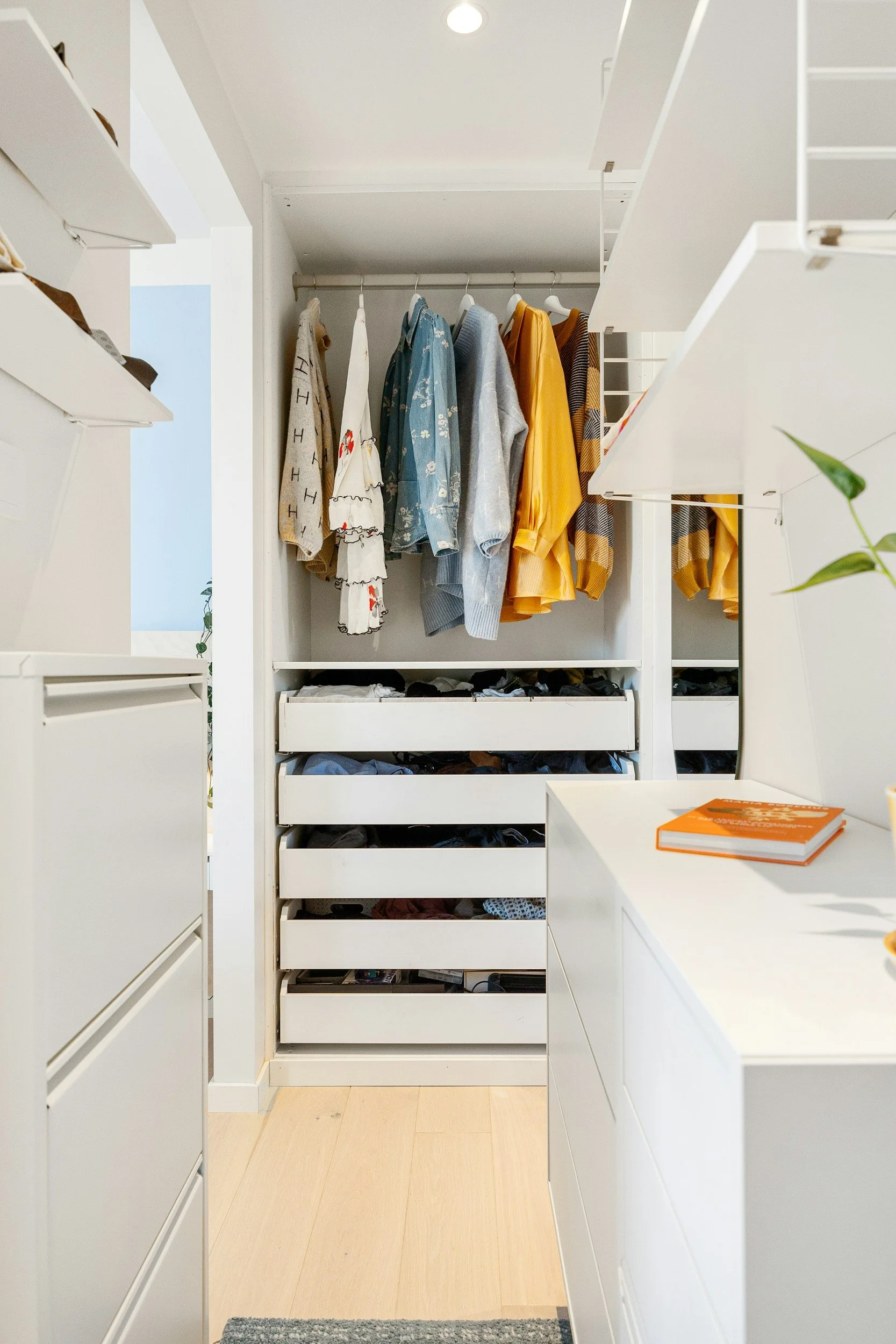 A walk-in closet with white shelves, drawers, and a hanging rod filled with colorful clothes. There is a small orange book on a white surface and a gray and white rug on a light wooden floor.