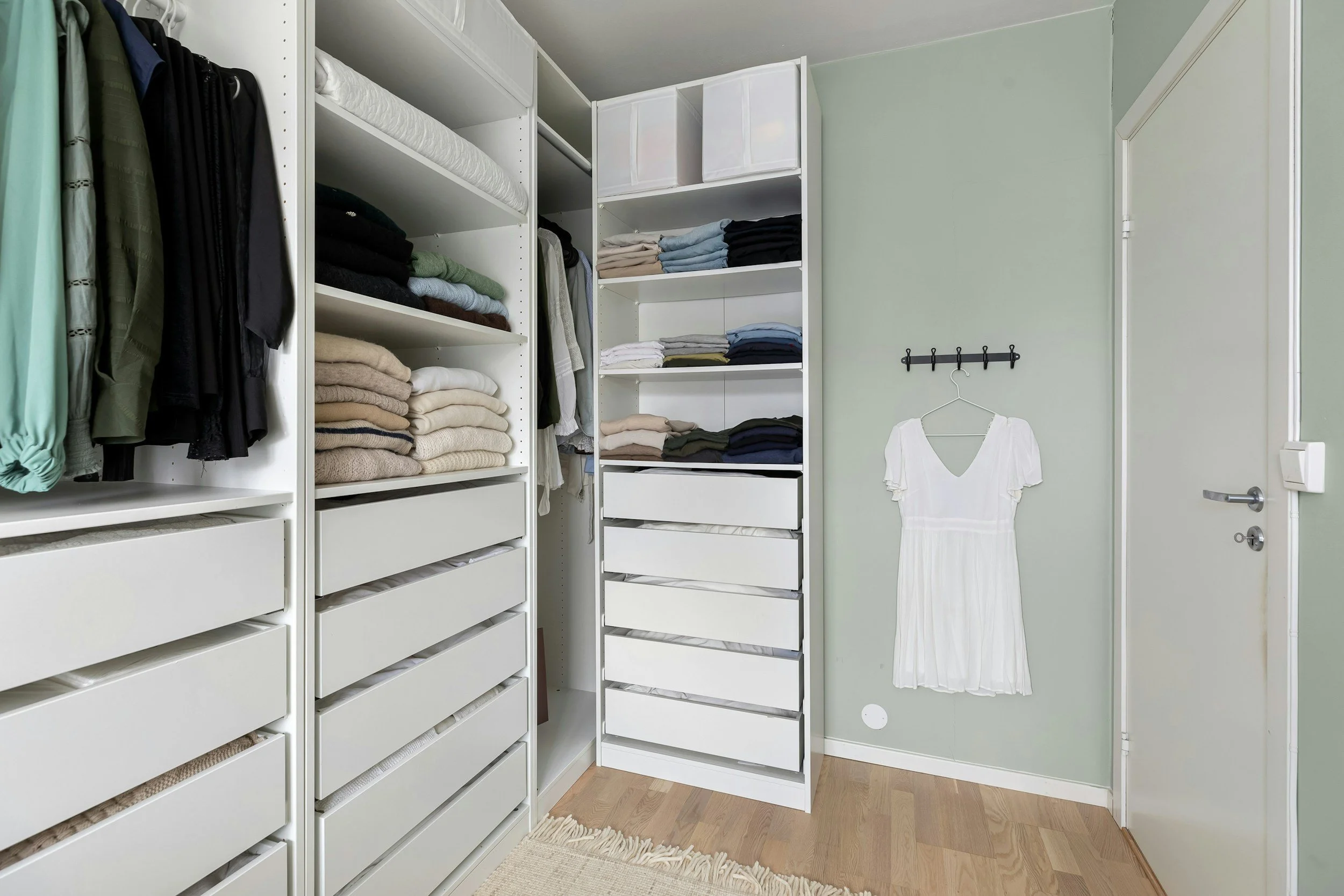 An organized walk-in closet with white shelving units filled with folded clothes, hanging garments, and storage boxes. A white dress hangs on a wall hook next to a door.