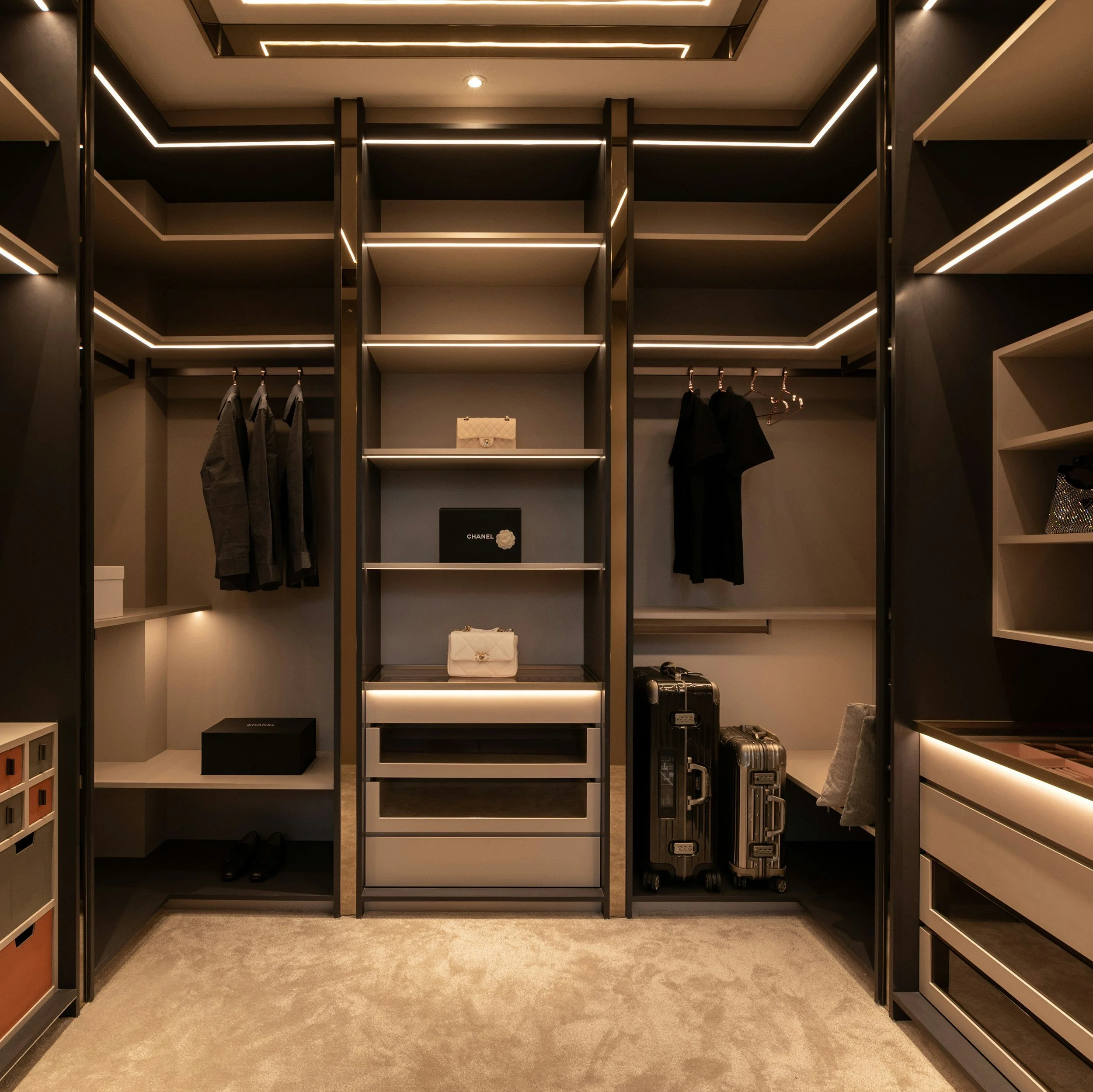 Walk-in closet with black shelving and clothing hanging, handbags on shelf, and luggage on the floor, illuminated with modern lighting.