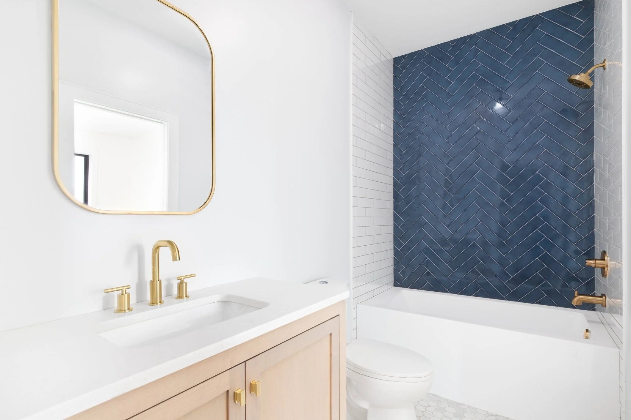 Modern bathroom featuring white vanity with gold fixtures, a large mirror with a gold frame, a white bathtub, and a shower area with dark blue herringbone tile walls. There are also smaller white tiles with a hexagonal pattern near the bathtub.