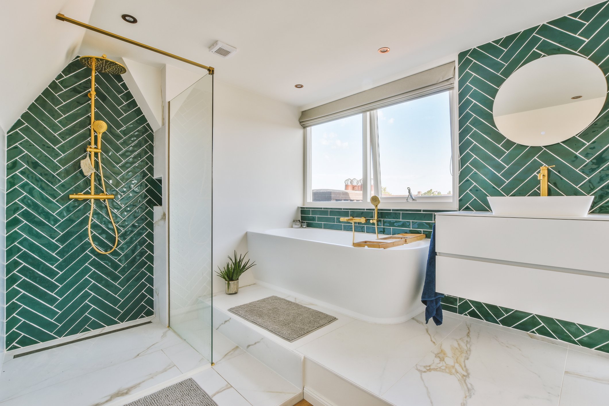 Modern bathroom with green tiled shower wall, gold fixtures, white bathtub, large window with blinds, round mirror, and marble floor.