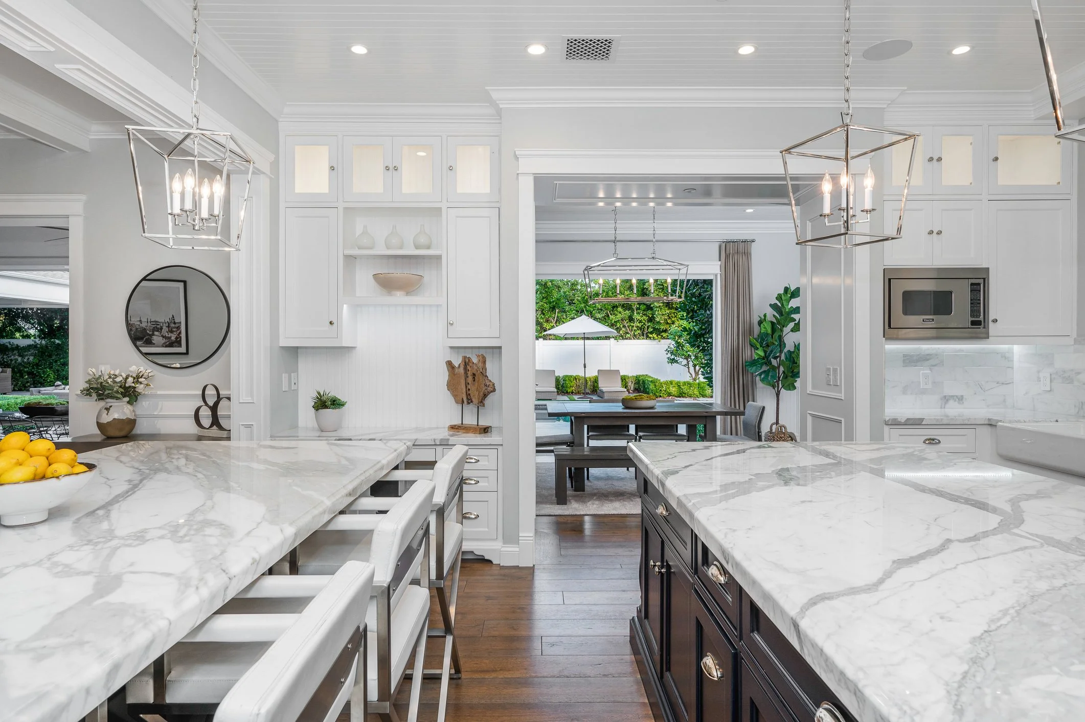 A modern kitchen with white marble countertops, white and dark wood cabinets, and stainless steel appliances. There are hanging pendant lights, decorative vases, a round mirror, and a window overlooking greenery, creating a bright and stylish space.