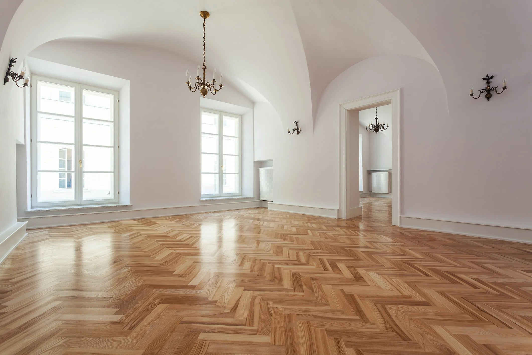 Spacious room with large windows, ornate chandelier hanging from ceiling, hardwood floor, white walls, and wall-mounted light fixtures.