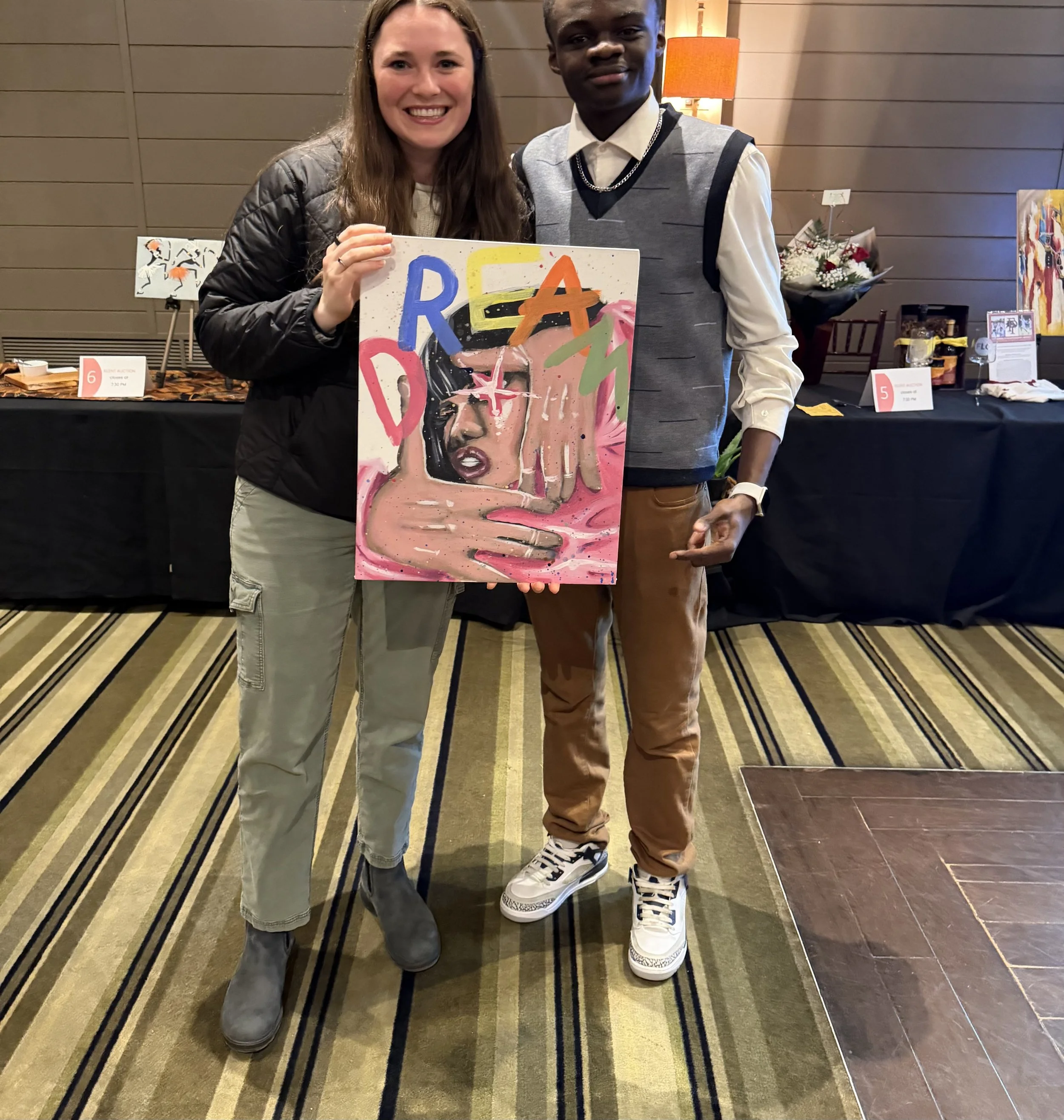 A woman and a young man standing together, smiling at the camera, holding a colorful painting of a woman. They are in an indoor space with a table behind them displaying various items and artwork.