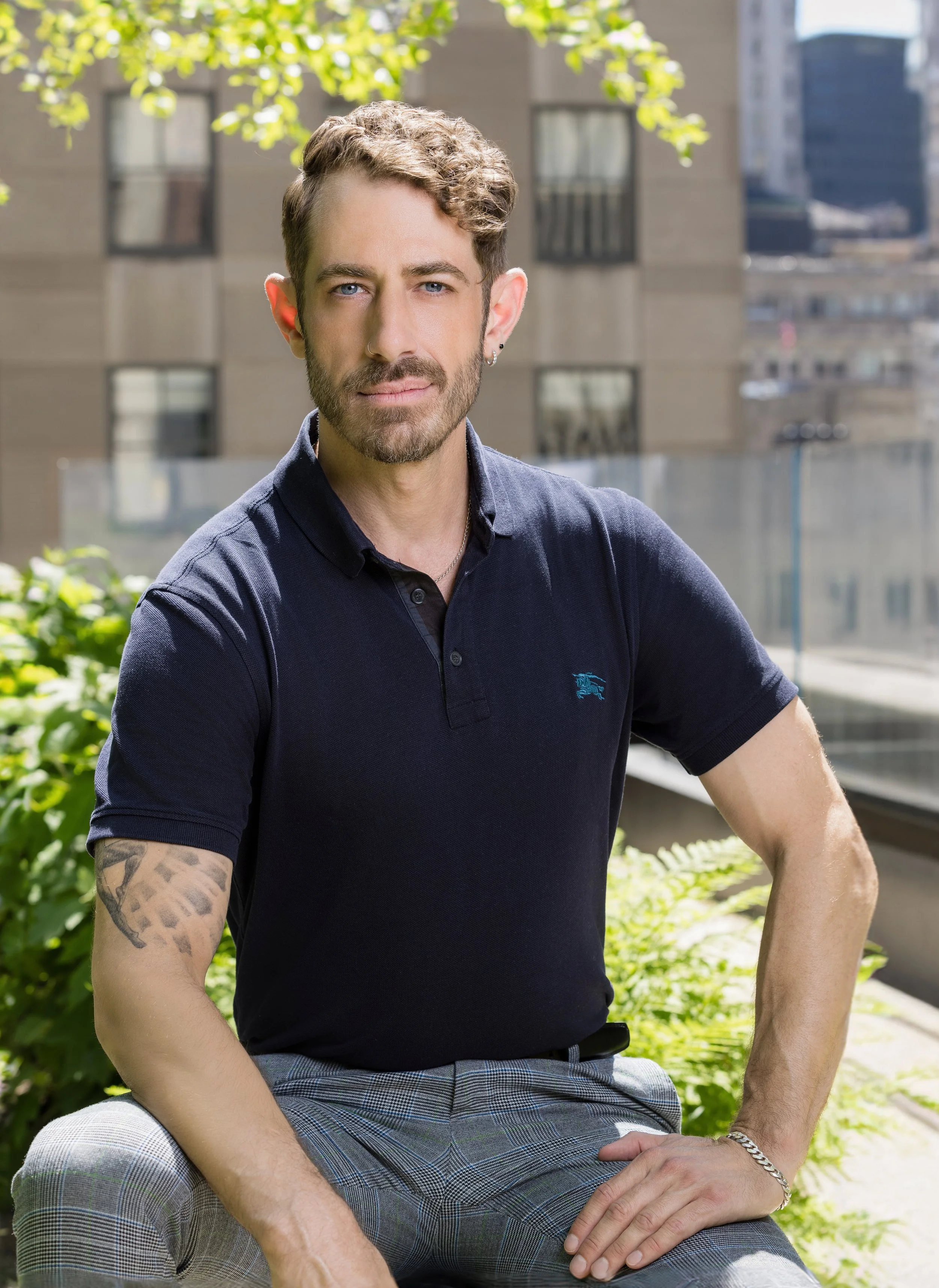 A young man with curly hair, a beard, and blue eyes, wearing a navy polo shirt and plaid pants, sitting outdoors on a sunny day with green trees and buildings in the background.