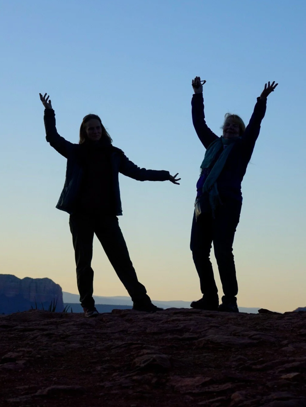 Two women in silhouette standing on rock at sunset with arms raised in front of a scenic landscape with cliffs and sky.