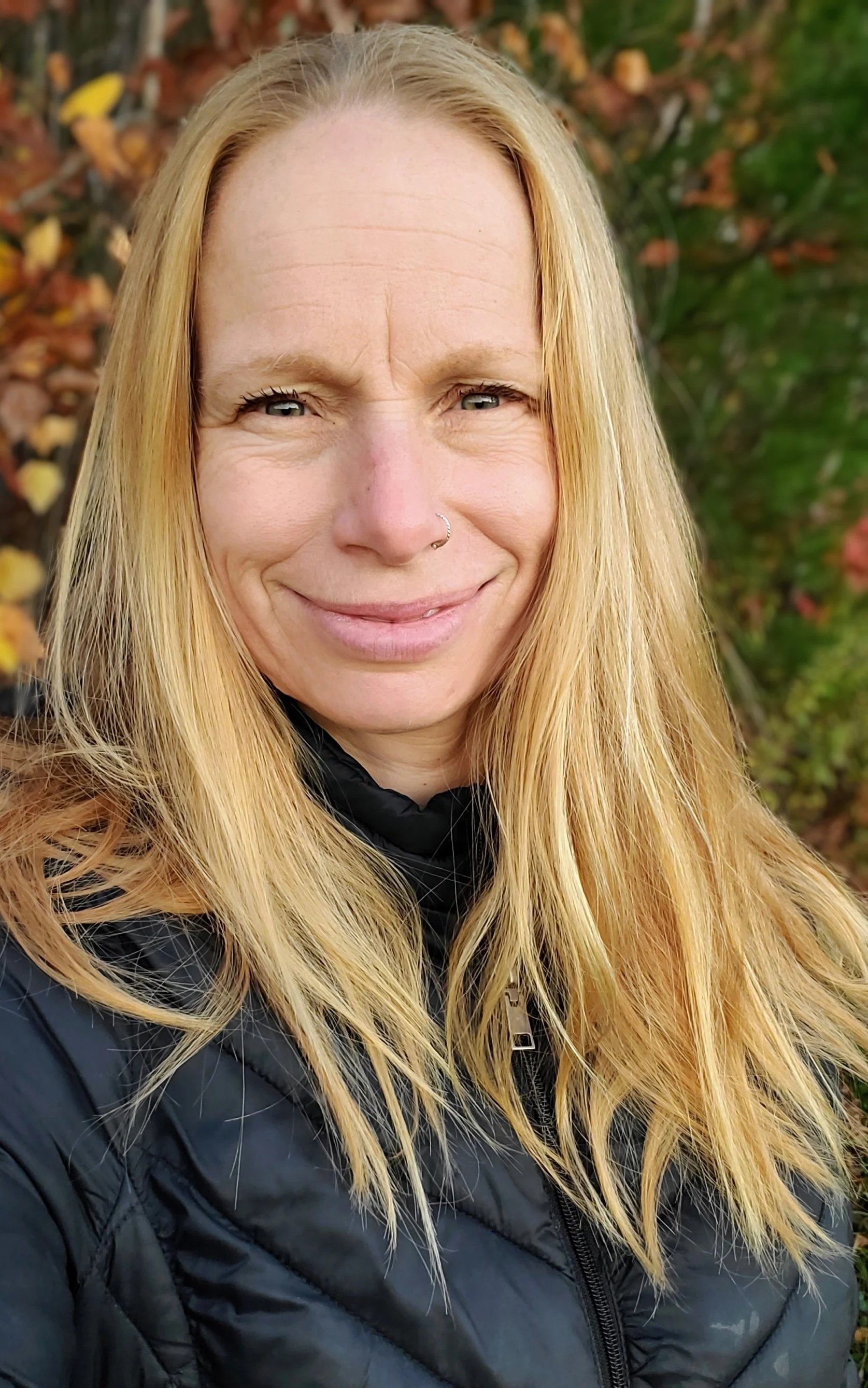 A woman with long blonde hair, wearing a black jacket with a zipper, standing outdoors with a background of autumn leaves and greenery.