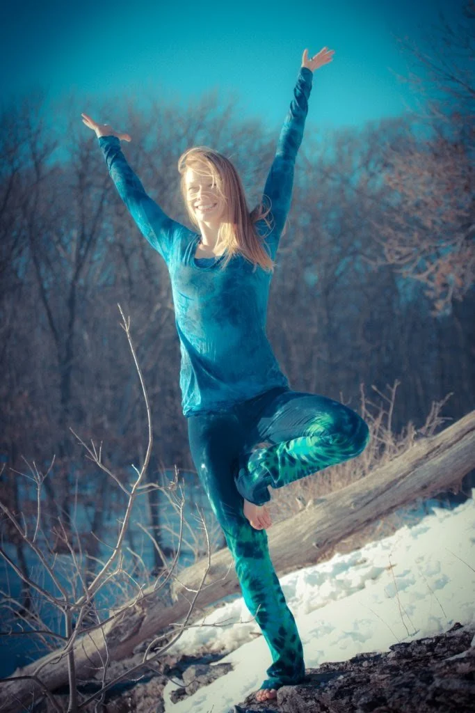 A young woman doing a yoga pose outdoors on snow-covered ground, with a fallen log and leafless trees in the background.