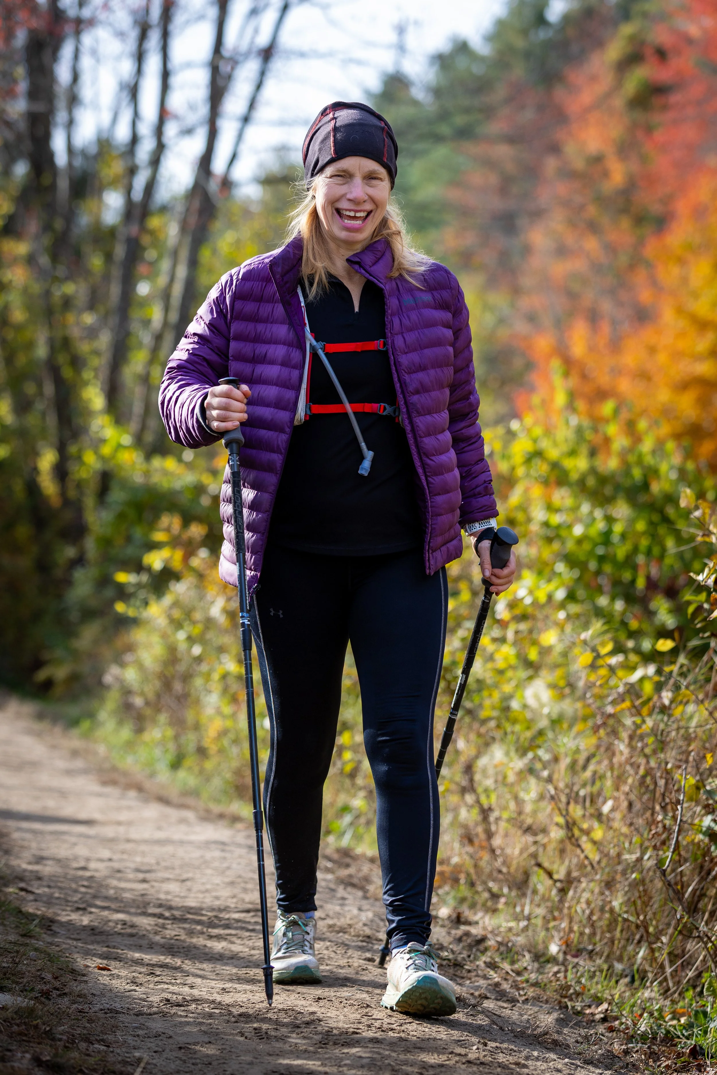 A woman hiking on a dirt trail through a wooded area with autumn-colored leaves, wearing a purple jacket, black pants, and a black hat, using trekking poles.