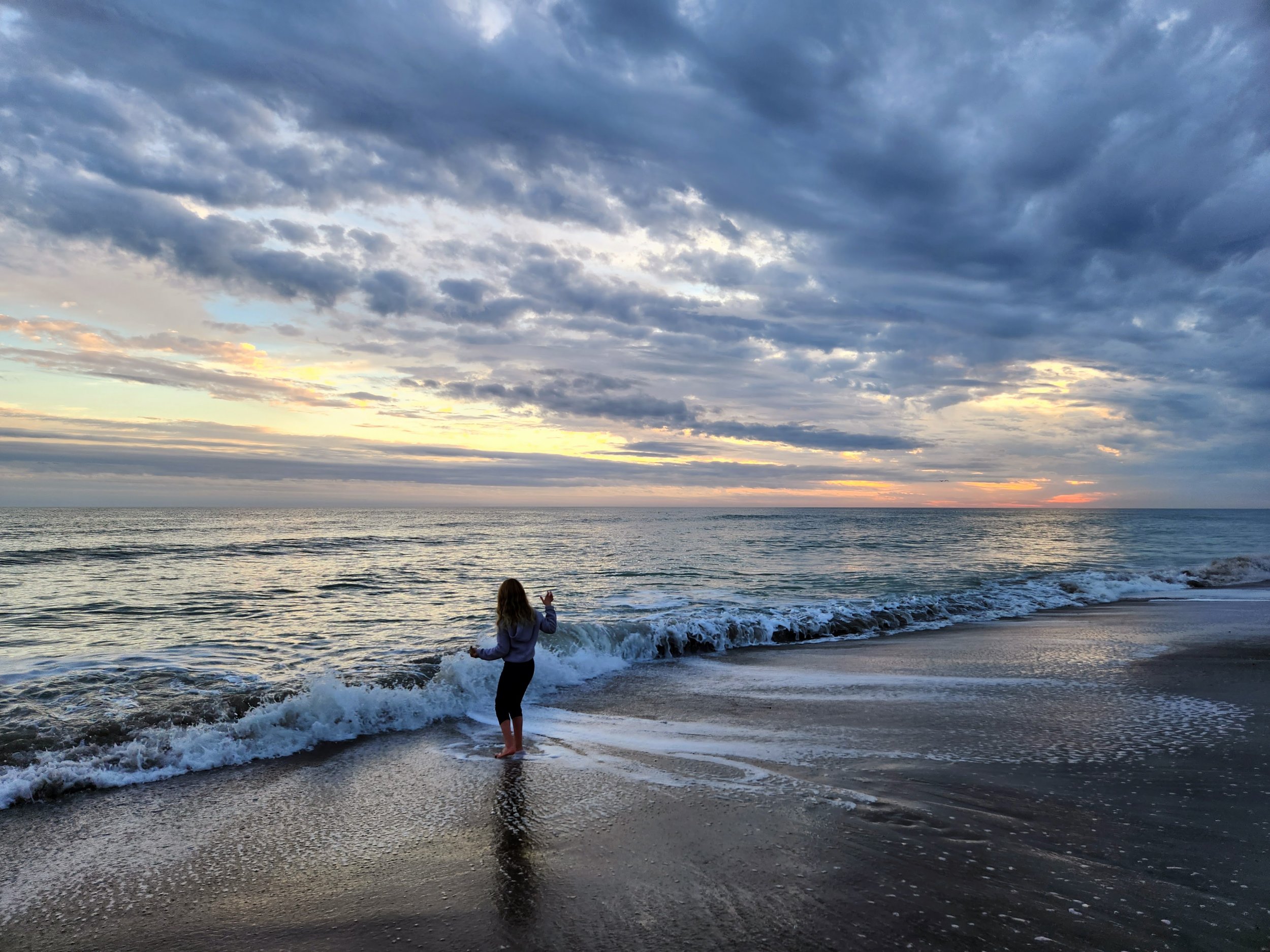 A person standing on a sandy beach as waves gently crash on the shore, with a sky filled with clouds during sunset or sunrise, creating a peaceful and scenic atmosphere.