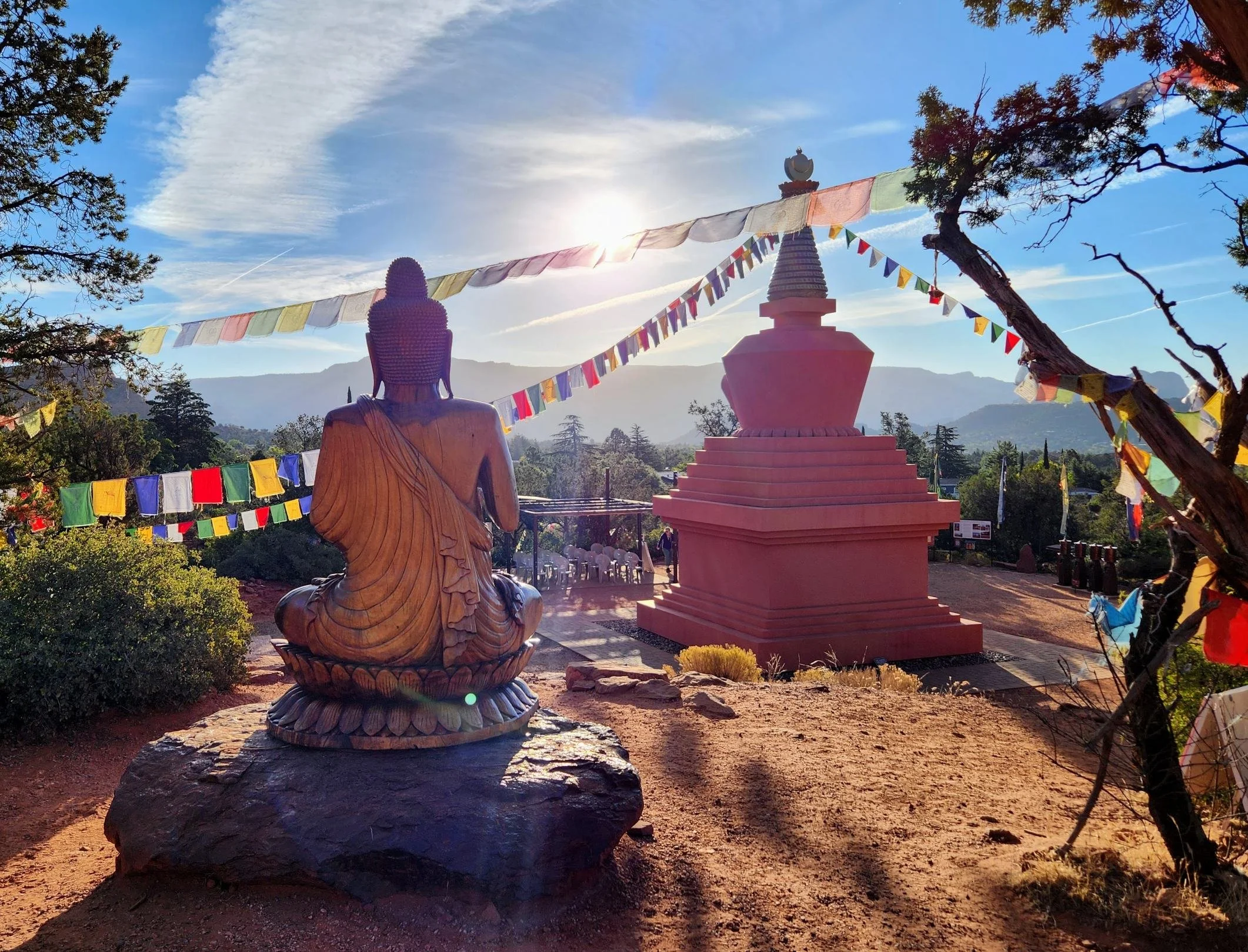 A Buddhist temple scene with a large bronze statue of Buddha seated on a rock in the foreground, and a stupa with prayer flags in the background, under a partly cloudy sky with the sun shining.