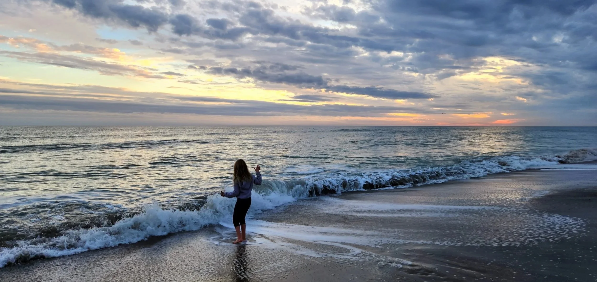 A person standing on a beach at sunset, looking at the ocean waves, with a partly cloudy sky overhead.