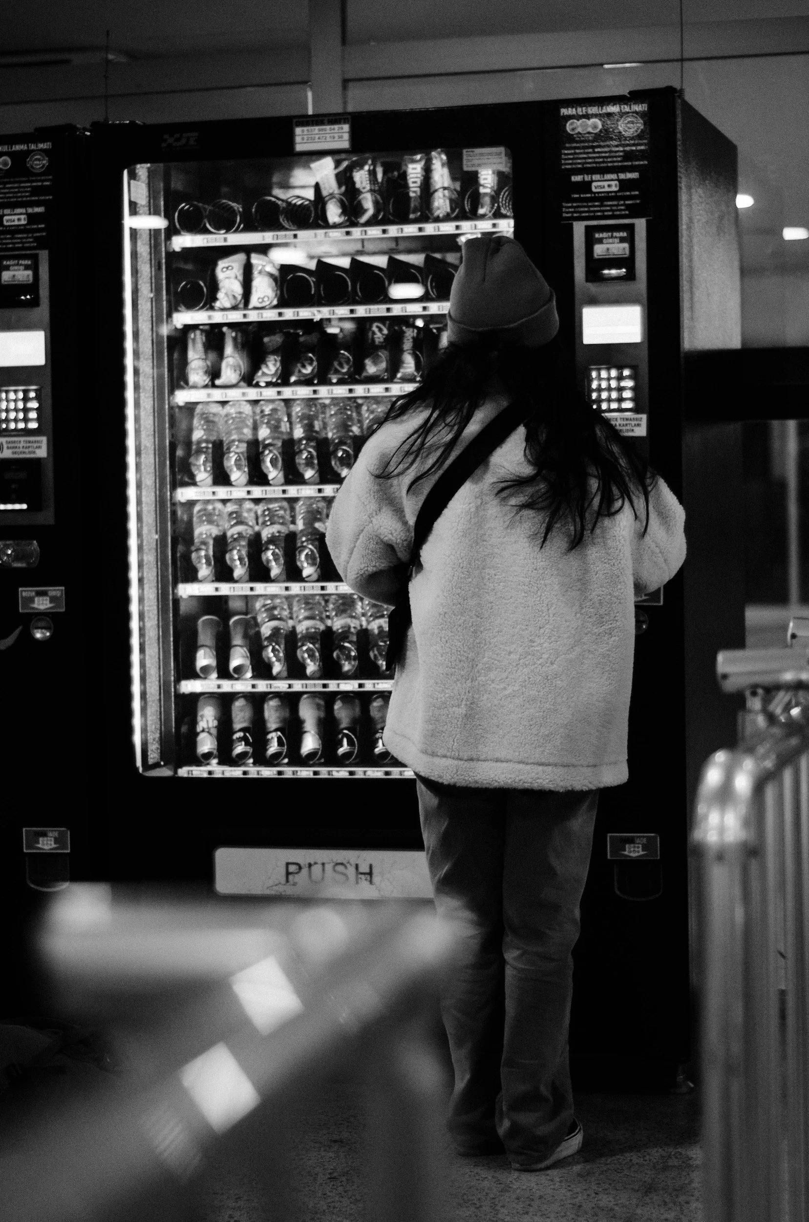 A person wearing a beanie and a coat standing in front of a vending machine with bottled drinks in an indoor setting, viewed from behind.