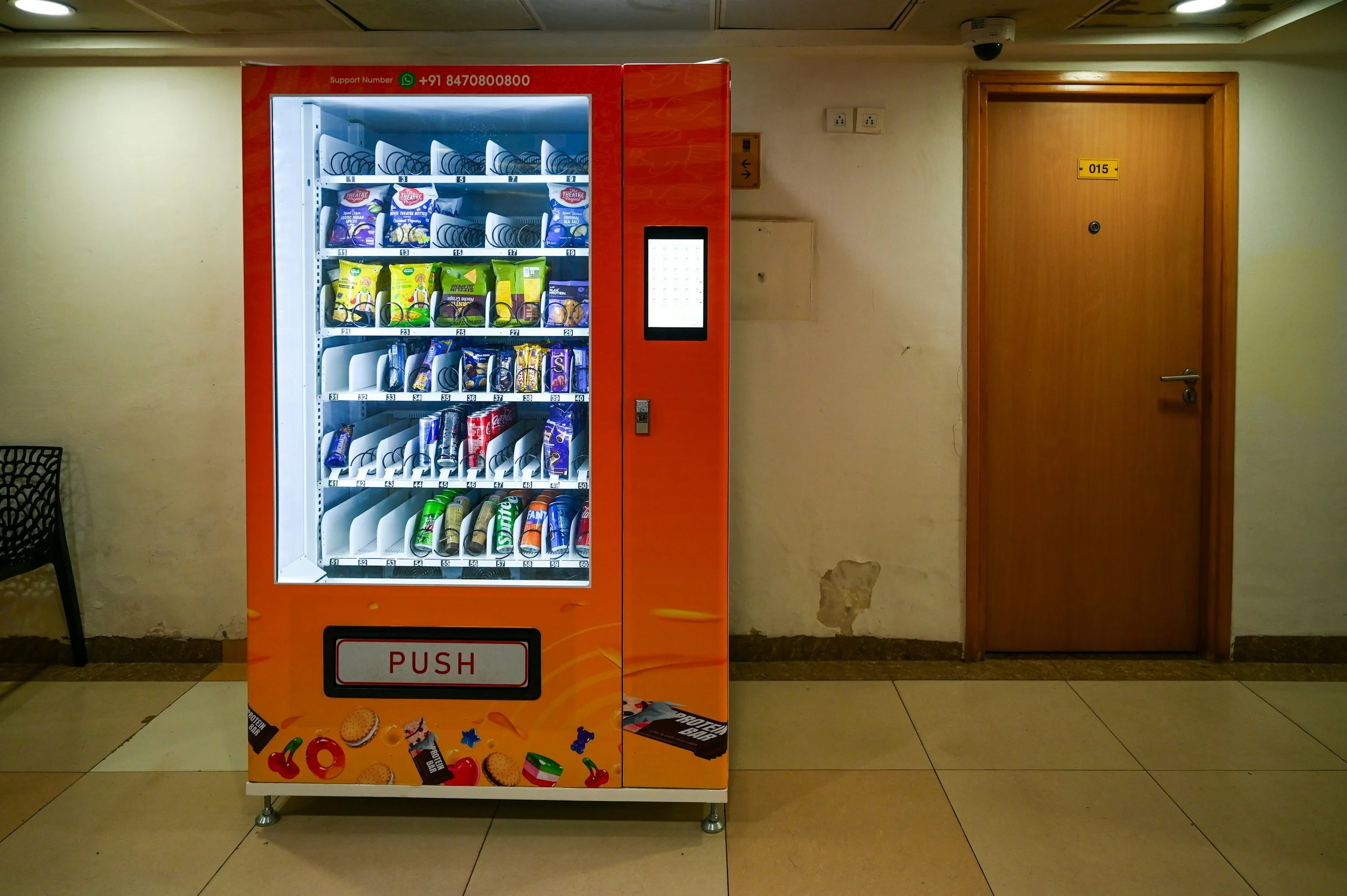A vending machine stocked with snacks is positioned against a beige wall with a wooden door to the right. The vending machine has a red and orange design with snack items visible inside. To the right of the vending machine are two electrical outlets on the wall.