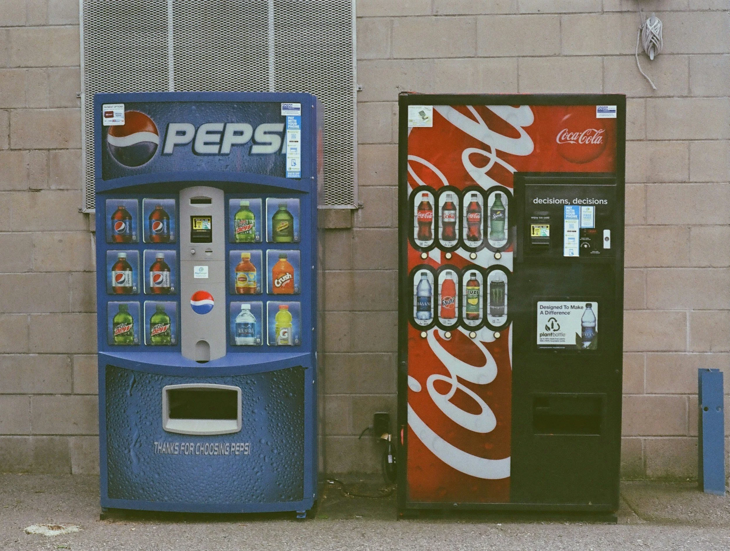 Two vending machines against a tan brick wall. The left machine is blue with a Pepsi logo, selling various beverages including Pepsi, Mountain Dew, Gatorade, and Crush. The right machine is black with a Coca-Cola logo, selling Coca-Cola, Sprite, bottled water, and energy drinks.