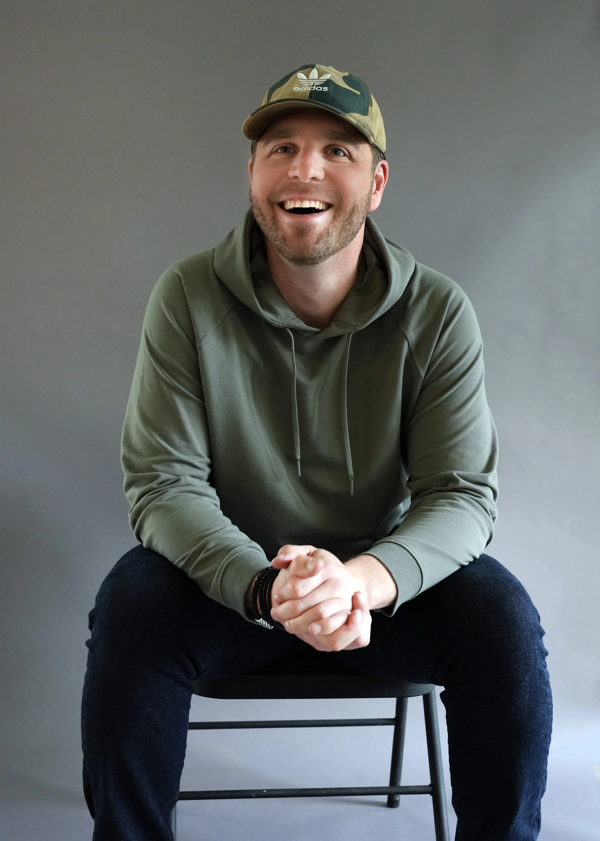 A man sitting on a black chair against a plain gray background, wearing a camouflage baseball cap, a green hoodie, and black pants, smiling and with his hands clasped together.