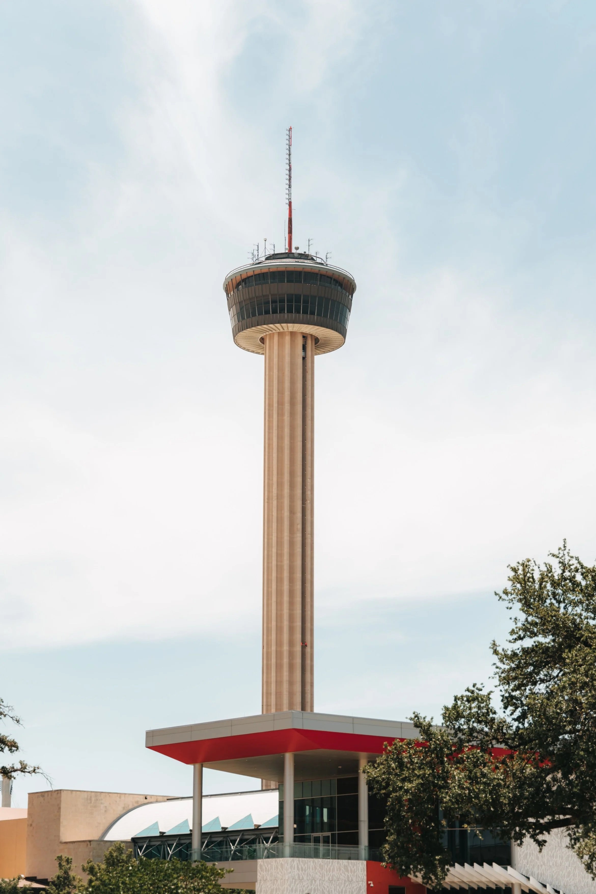 A tall building with a cylindrical observation deck and a red and white antenna on top, situated beside a modern structure with glass walls and a red roof edge, with trees in the foreground and a cloudy sky in the background.