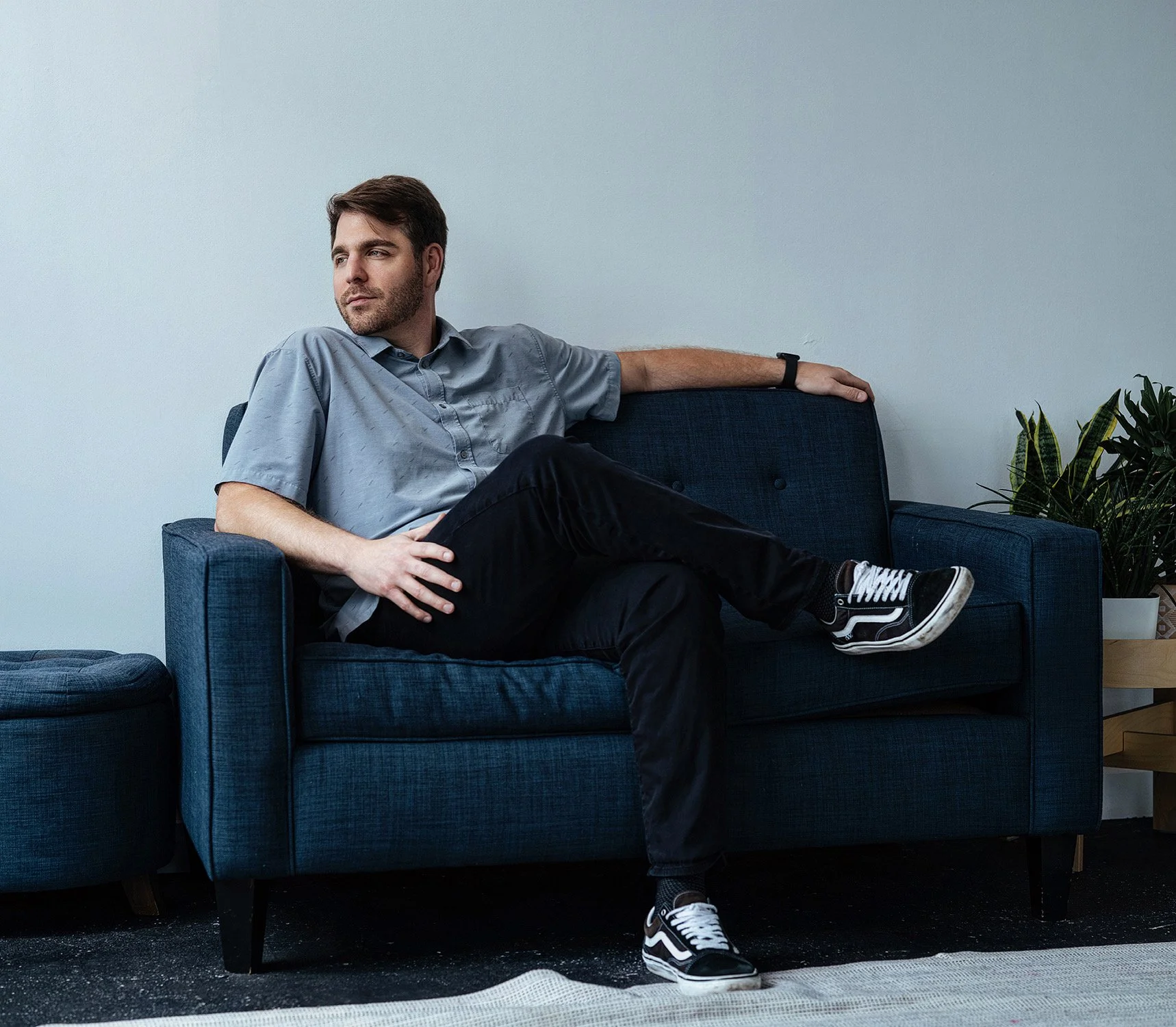 A man sitting on a blue sofa with his arm resting on the back, wearing a gray shirt, black pants, and sneakers, next to a potted plant.