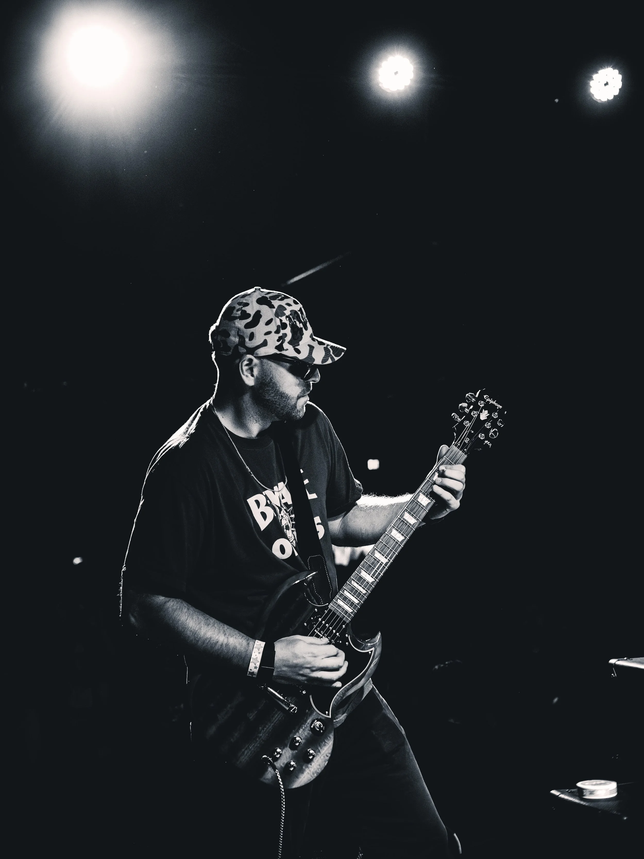 A man wearing sunglasses and a camouflage cap playing an electric guitar on stage, with bright spotlights overhead.