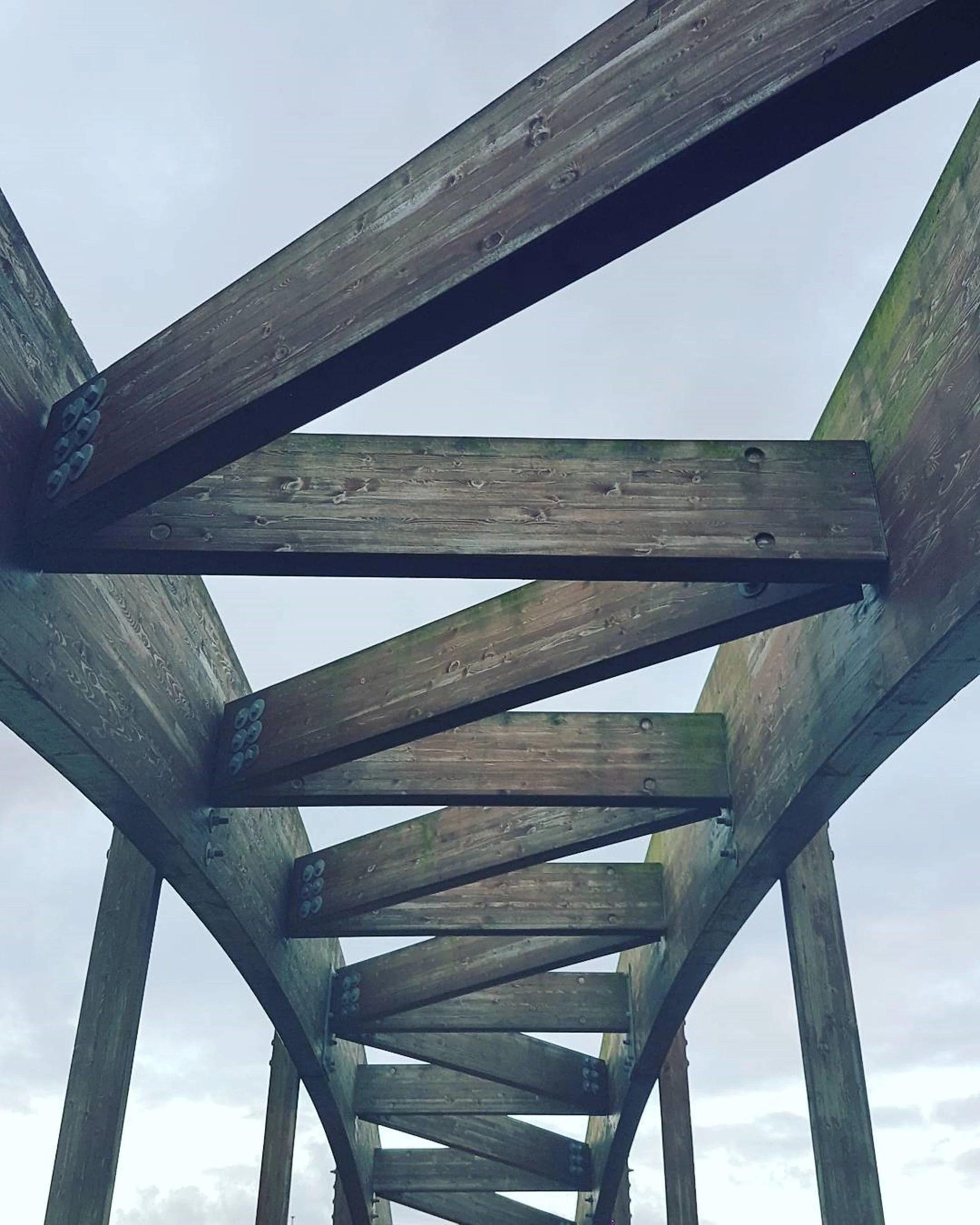 Upward view of wooden structure, possibly a bridge or platform, with support beams and bolts against a cloudy sky.