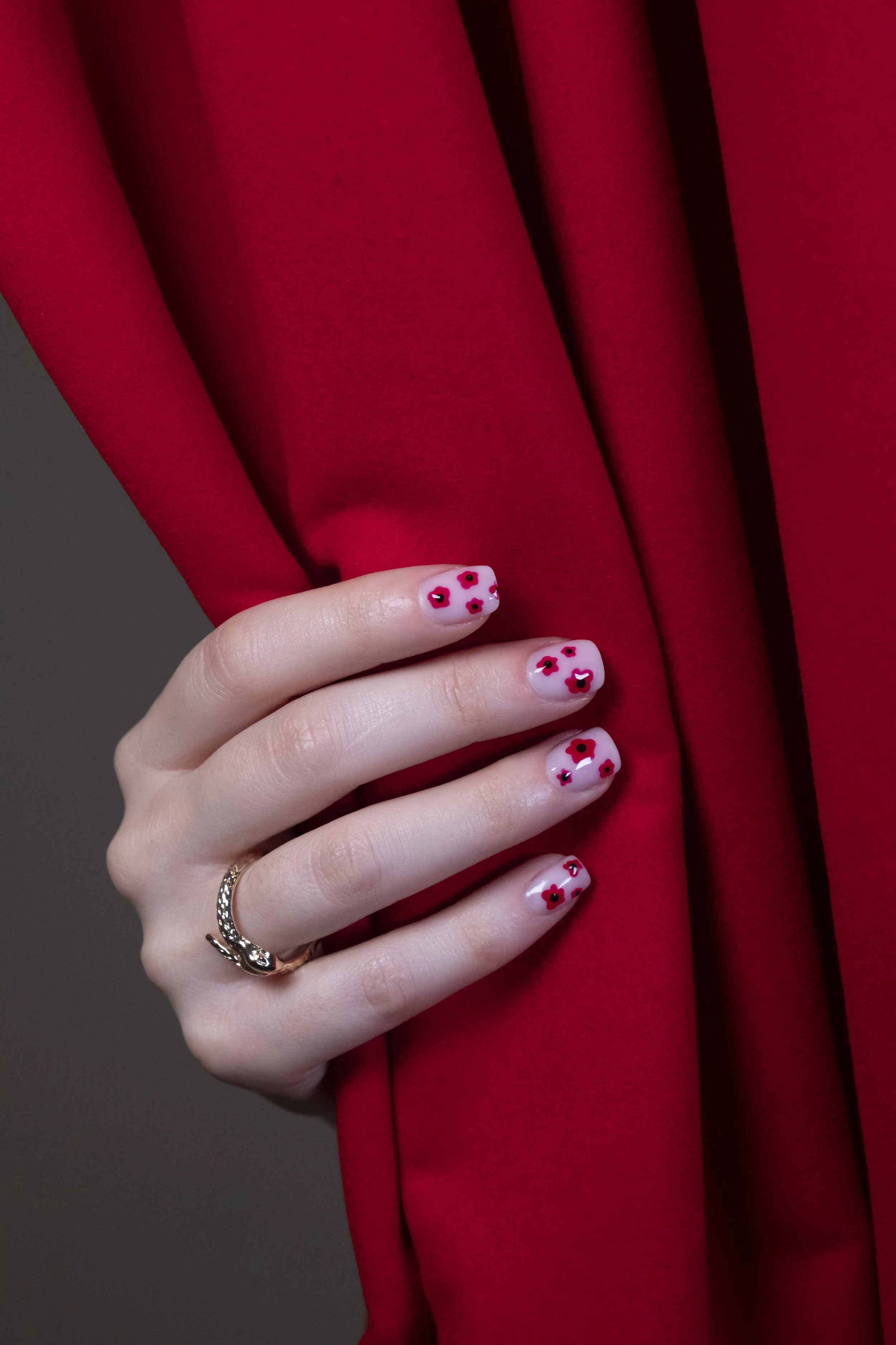 Una mano con un anello d'argento indossa unghie con smalto trasparente e decorazioni di fiori rossi. La mano si appoggia su un tessuto rosso.