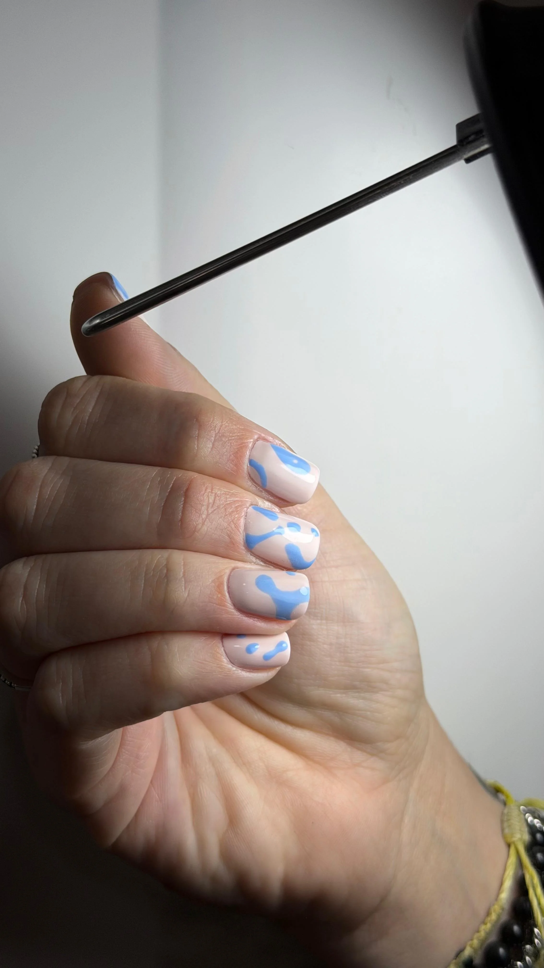 Mano con unghie decorate con smalto bianco e motivi a onde blu chiaro, mentre tiene un bastone o un attrezzo nero in mano.