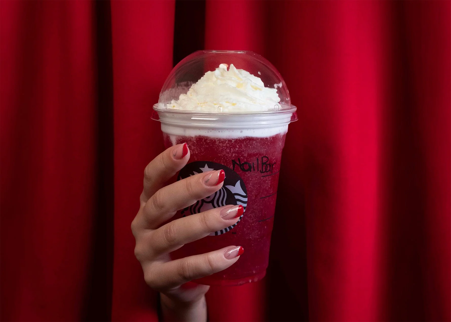 Una mano con unghie dipinte di rosso e bianco tiene una bevanda rossa con panna montata sopra, con uno sfondo di tende rosse.